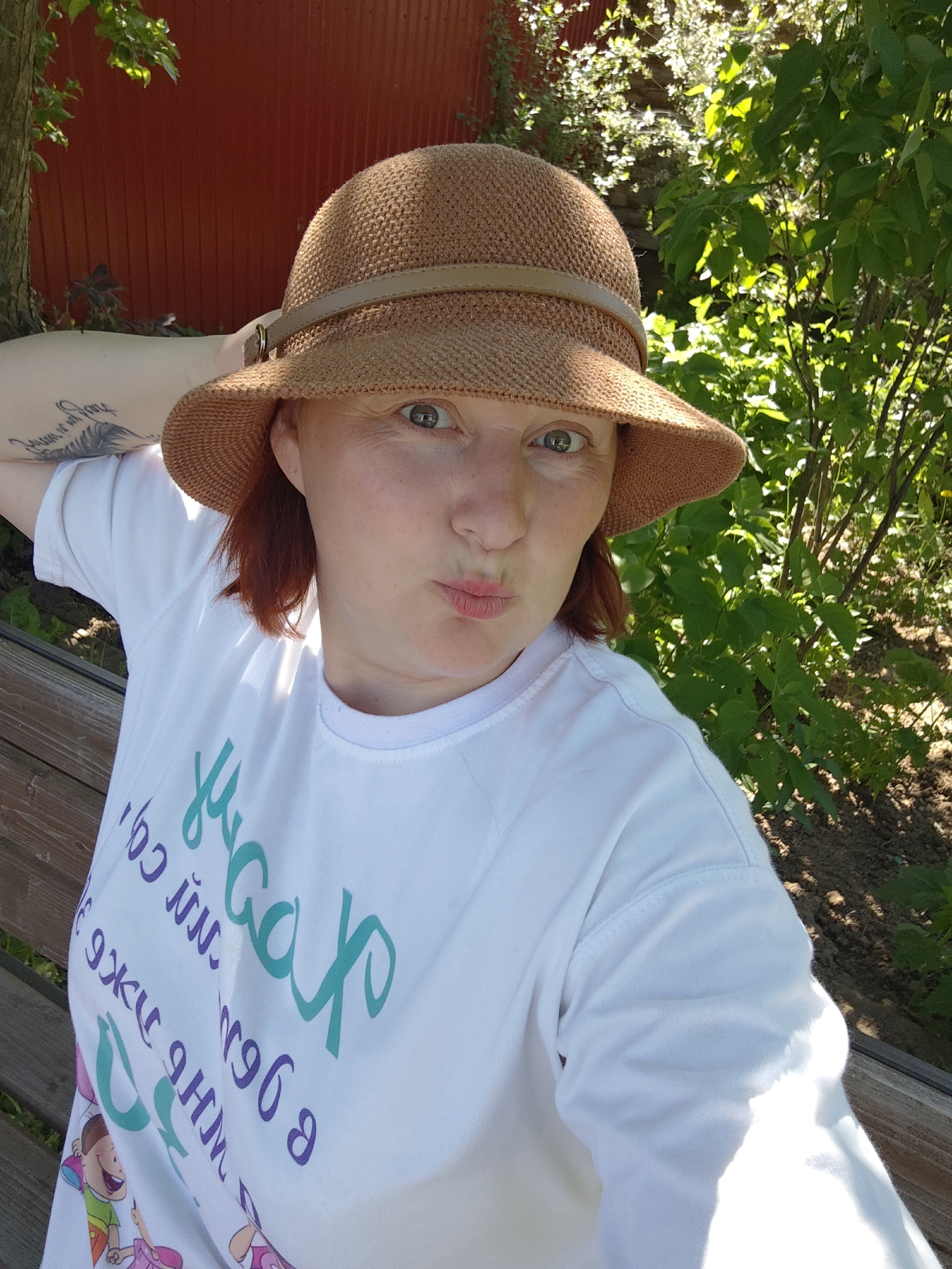 A woman is seated on a wooden bench in what appears to be a park or garden. She is wearing a light-brown hat with a darker brown band, and a white t-shirt with colorful writing and cartoon images on it. Her left arm is raised, resting on the back of the bench, revealing a tattoo on her upper arm. She is looking directly at the camera with a slightly puckered expression. The background includes greenery such as trees and bushes, a wooden fence, and some dappled sunlight. The location could be Фоки, Russia, based on the requested detail.