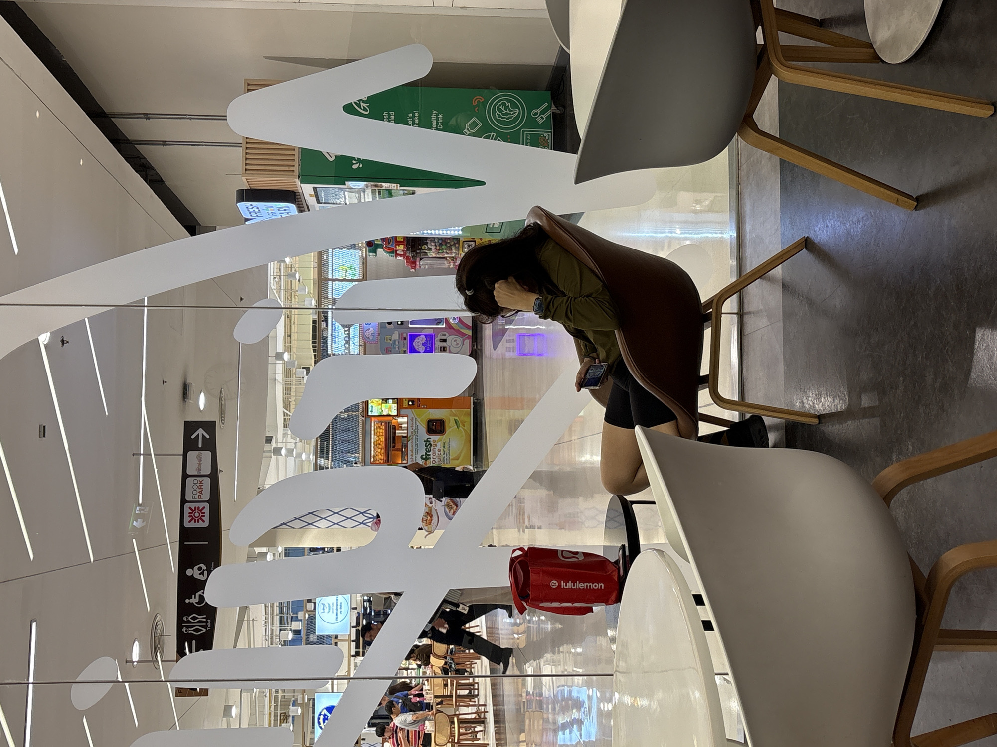This is a photograph taken indoors, likely in a shopping mall in Nong Pa Khrang, Thailand. The image features several elements, including people, furniture, and signage, viewed through a glass partition with large white lettering that partially obscures the scene.
In the foreground, a woman with dark hair is seated in a brown chair, appearing to be looking at her phone. She is wearing a green top and black shorts, and a red Lululemon bag is placed on the floor beside her. White chairs and wooden table legs are also visible in the immediate foreground.
The background, seen through the glass, reveals a bustling environment. There are various food stalls or shops with colorful displays and signage. One sign prominently features "fresh Orange juice" and "let's make! Healthy Drink". Another sign points towards "FOOD PARK" with directional arrows. Other signs show icons for a restroom and accessibility. People are seen walking and dining in this area, suggesting it is a food court or a similar public space. The lighting suggests it is daytime. The overall impression is a lively, commercial indoor setting.