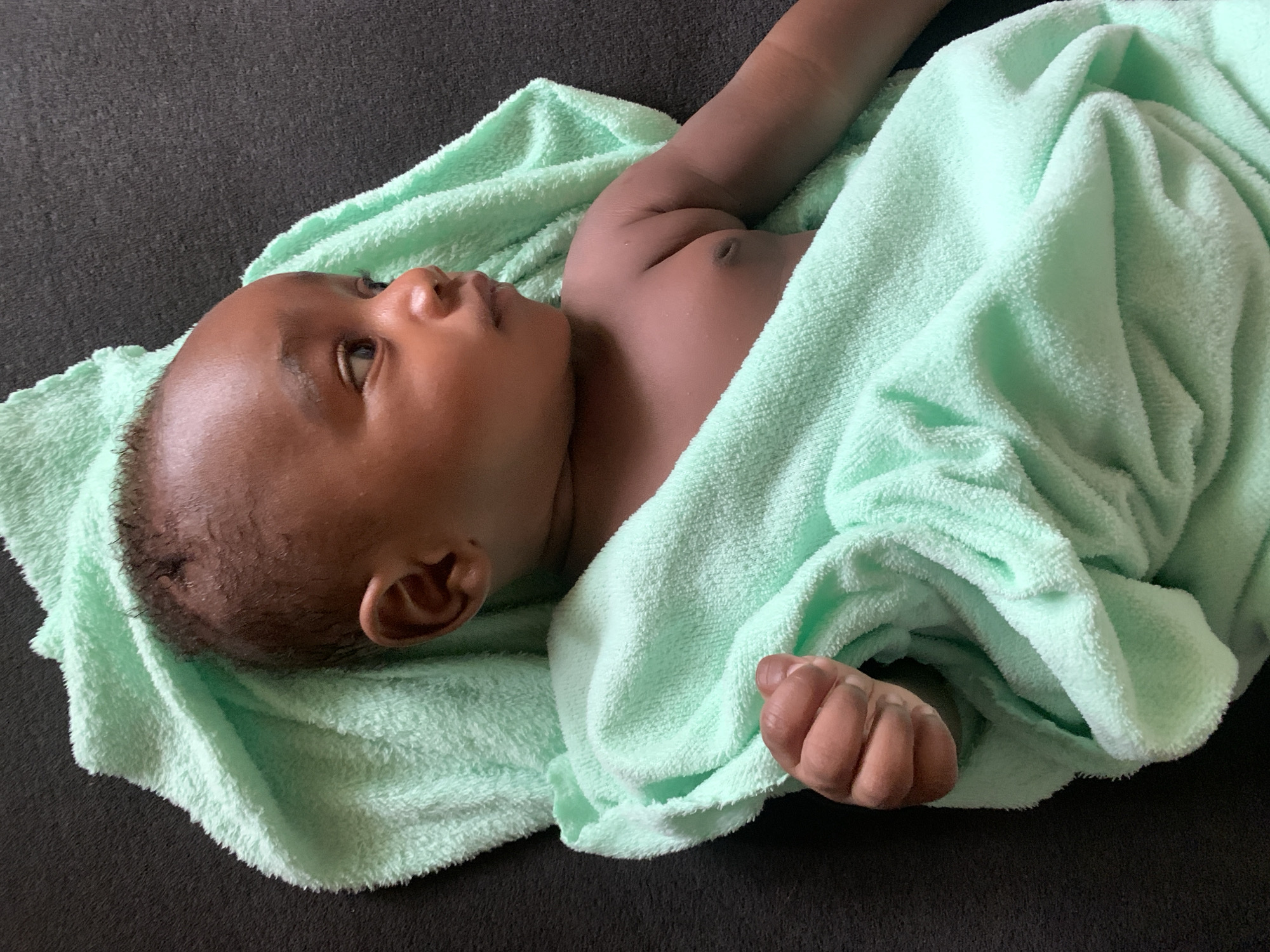 A baby lies on a dark surface partially wrapped in a light green towel. The baby's head is towards the left of the frame, and the body angles towards the upper right. The baby is dark-skinned and is looking towards the left with eyes open. The location is unspecified, and there is no indication that the image was taken in Barakin Akawo, Nigeria.