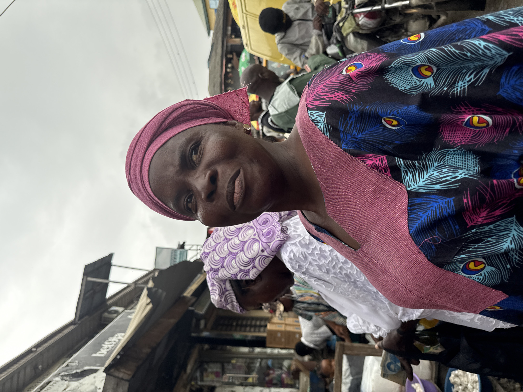 In the city of Ado, Nigeria, a woman is captured in a close-up portrait, her face turned slightly to the right. She wears a vibrant pink headscarf, a pearl earring, and a colorful dress adorned with a peacock feather pattern. Behind her, a bustling marketplace unfolds under a cloudy sky. Another woman in a purple and white patterned headscarf and a white garment stands to her left. In the background, men are visible, some near motorcycles, suggesting the presence of transportation common in Nigerian markets. The overall scene depicts the lively atmosphere of a typical Nigerian urban market.