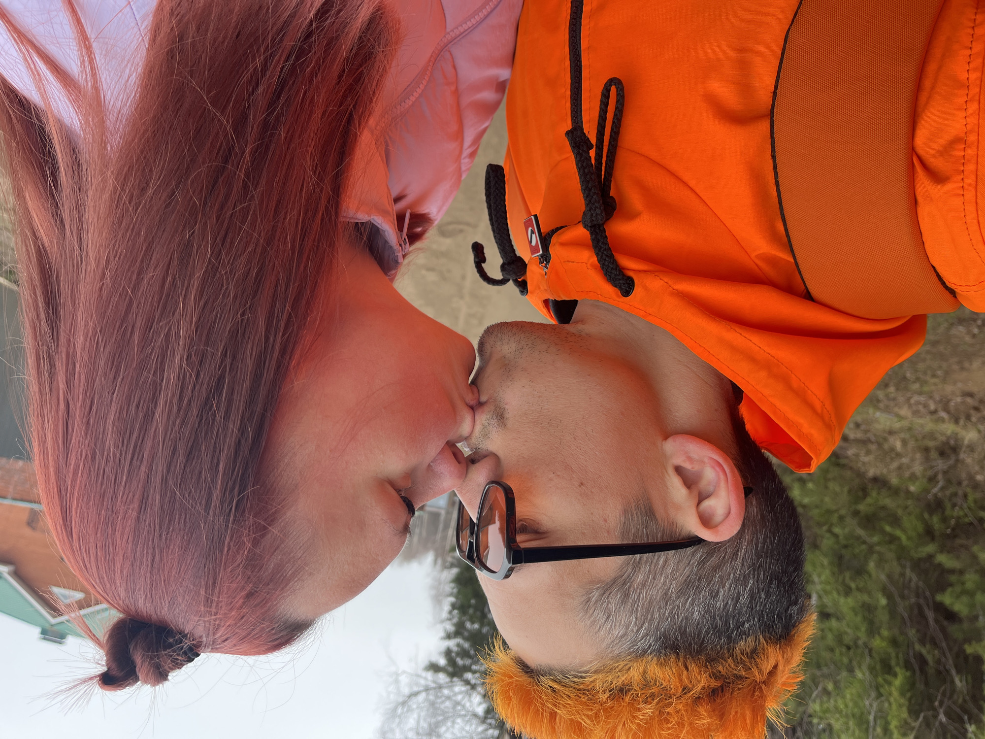This vertically oriented image captures two individuals, a man and a woman, kissing outdoors in Chaikovsky, Russia. The woman, positioned on the left, has long reddish-pink hair and wears a light pink zippered jacket. The man, on the right, has short gray hair and is wearing a vibrant orange jacket with black drawstrings. A distinctive orange furry trim adorns his neck or hood area, and he wears glasses with black frames and tinted lenses. Their lips are touching in a tender kiss, conveying affection and intimacy.
The background is muted and slightly out of focus, indicating an overcast or hazy daytime setting. Bare trees and what appears to be a distant building with a green roof are subtly visible, suggesting a natural or semi-urban outdoor environment, possibly during early spring or late autumn. A small, unreadable red and white logo is present on the man's orange jacket. The overall atmosphere is one of a private and loving moment shared between the couple.