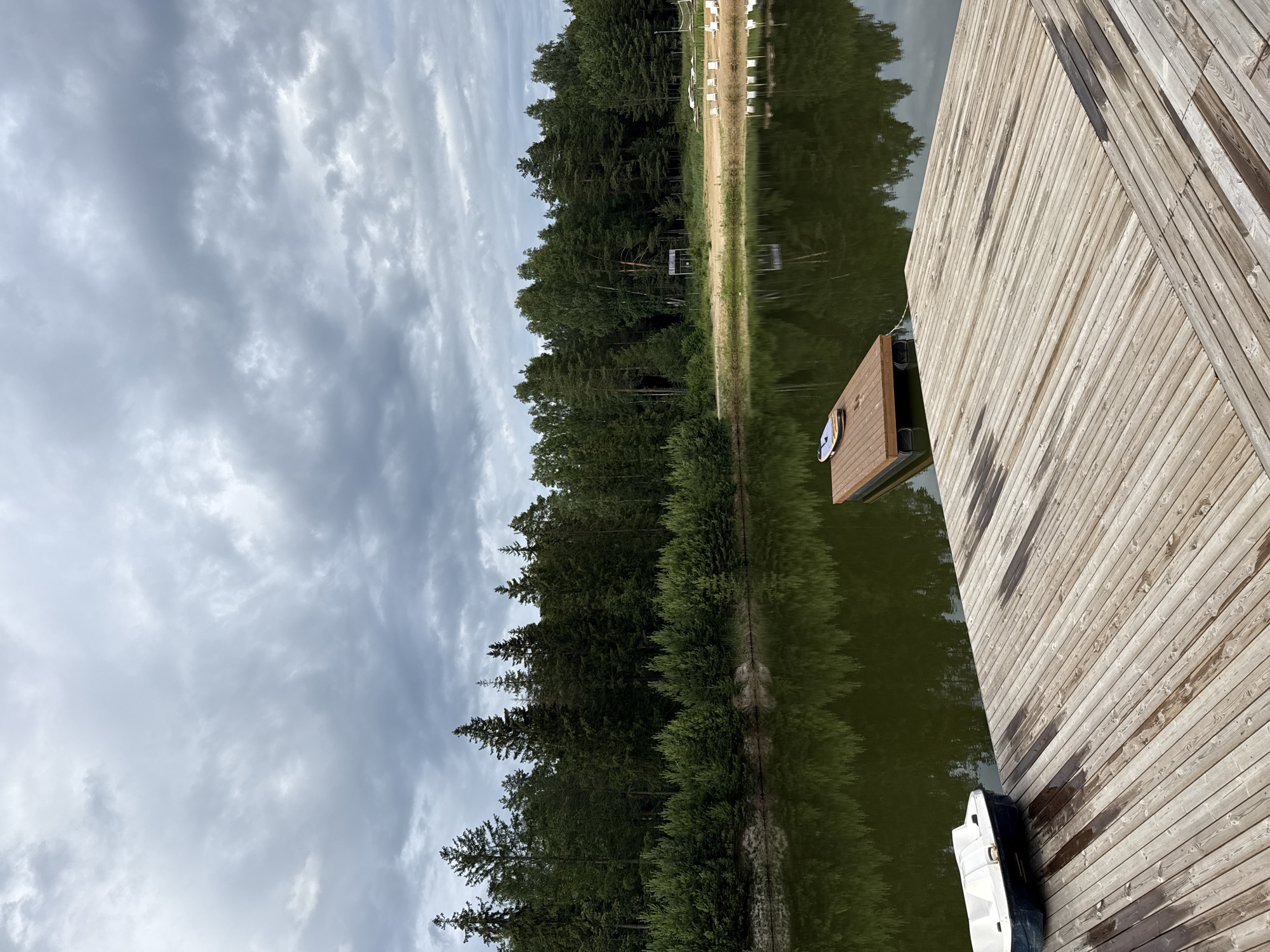 The image depicts a serene lake scene with a focus on a wooden dock and a surrounding natural landscape. The dock, constructed from weathered wooden planks, occupies the right side of the frame and extends into the water. A small, dark-colored object, possibly a bag or a case, rests on the dock near its edge. A white boat is moored to the dock, partially submerged in the calm, dark water.
The lake itself is a prominent feature, its surface reflecting the dense green forest that lines the opposite shore. The trees are a mix of tall pines and other deciduous species, creating a lush and verdant backdrop. A narrow strip of sandy beach is visible between the water's edge and the treeline. Beyond the beach, more trees and some white structures, possibly beach chairs or changing booths, are faintly visible.
The sky above is overcast with a layer of textured gray clouds, suggesting a temperate day, possibly late afternoon or early morning given the soft, diffused light. The overall mood is tranquil and peaceful, emphasizing the natural beauty of the setting. There is no visible text within the image. The location is identified as Sosnovka, Russia.