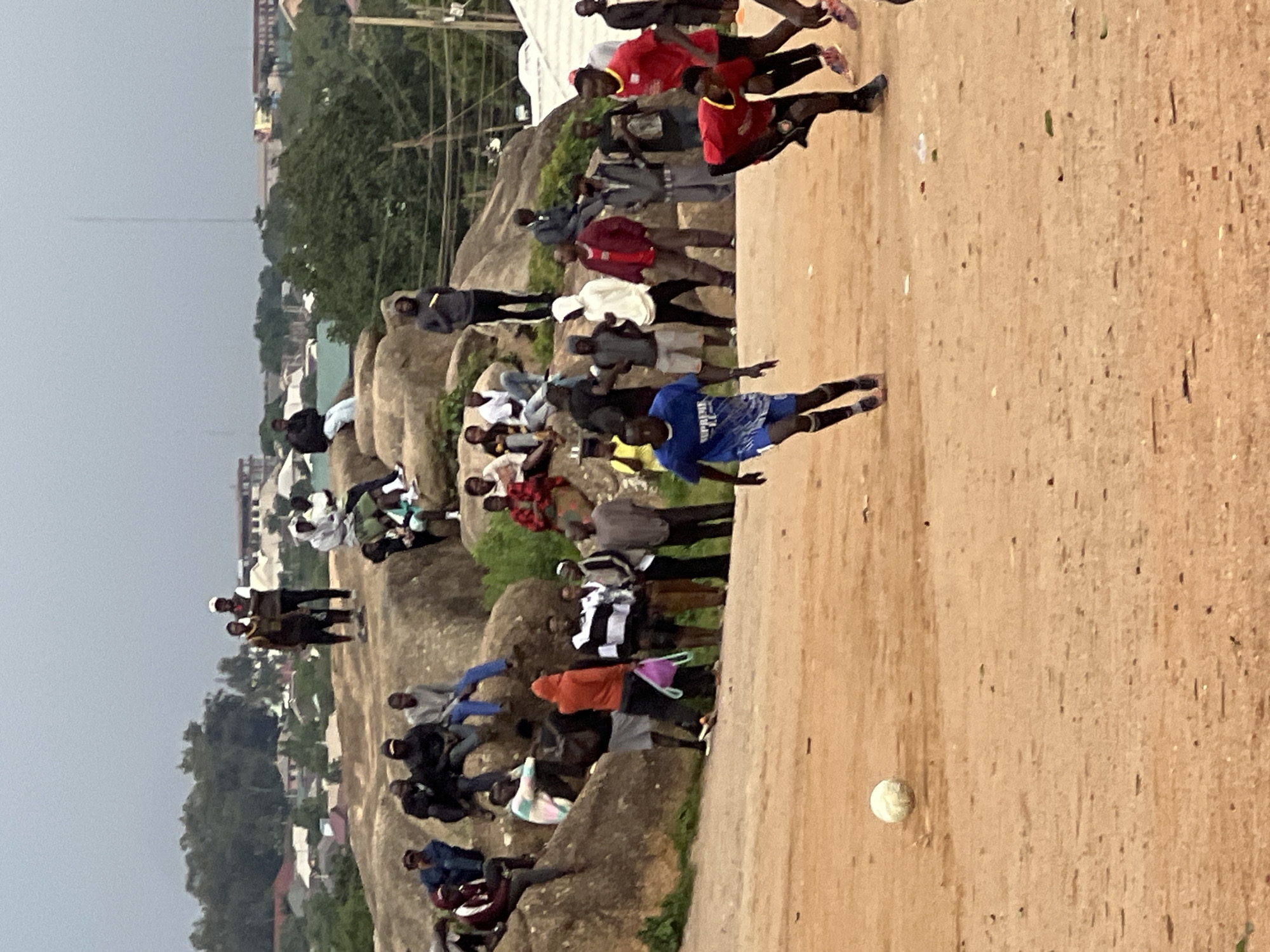 The image depicts an outdoor scene in Jos, Nigeria, likely a public gathering or informal sporting event. A group of people, predominantly young men and boys, are gathered in a rocky, uneven terrain. The main activity appears to be a football game, with one person in the foreground wearing a blue jersey and shorts, seemingly engaged in play. Several other individuals are scattered around, some watching the game, others standing or sitting on the large rocks that dominate the landscape. The atmosphere seems casual and engaged, with spectators observing the action.
The setting is characterized by a mix of natural and man-made elements. Large, weathered rocks form a significant part of the environment, providing natural seating and vantage points for the onlookers. In the background, a cityscape is visible, suggesting the location is on the outskirts or within a populated area. Buildings, trees, and possibly infrastructure like power lines can be seen, indicating an urban or semi-urban setting. The weather appears overcast, with a muted sky, suggesting it might be late afternoon or an overcast day. There is a single football visible on the sandy ground.
The people are dressed in various casual attire, including t-shirts, shorts, and some jerseys, consistent with a sporting event. Some individuals are wearing headwear. The overall impression is one of community involvement and engagement in a local pastime. No specific text is clearly legible within the image, though the blue jersey has some faint markings that might be text or a logo, but they are not discernible.