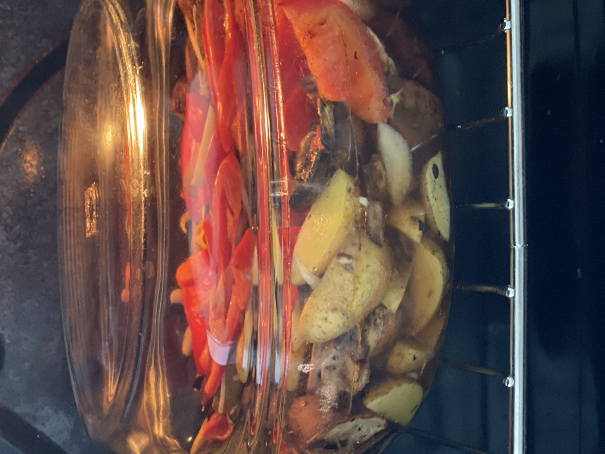 A close-up, eye-level shot shows a clear glass pot filled with food being cooked inside an oven. The pot contains sliced potatoes, tomatoes, and what appear to be onions, all submerged in liquid. The lid of the pot is slightly ajar, revealing the steam and cooking contents. The oven rack is visible beneath the pot, with small droplets of liquid clinging to its surface, suggesting the cooking process is underway. The background is the dark interior of the oven. There is no information available about the location of this scene in Solingen, Germany.