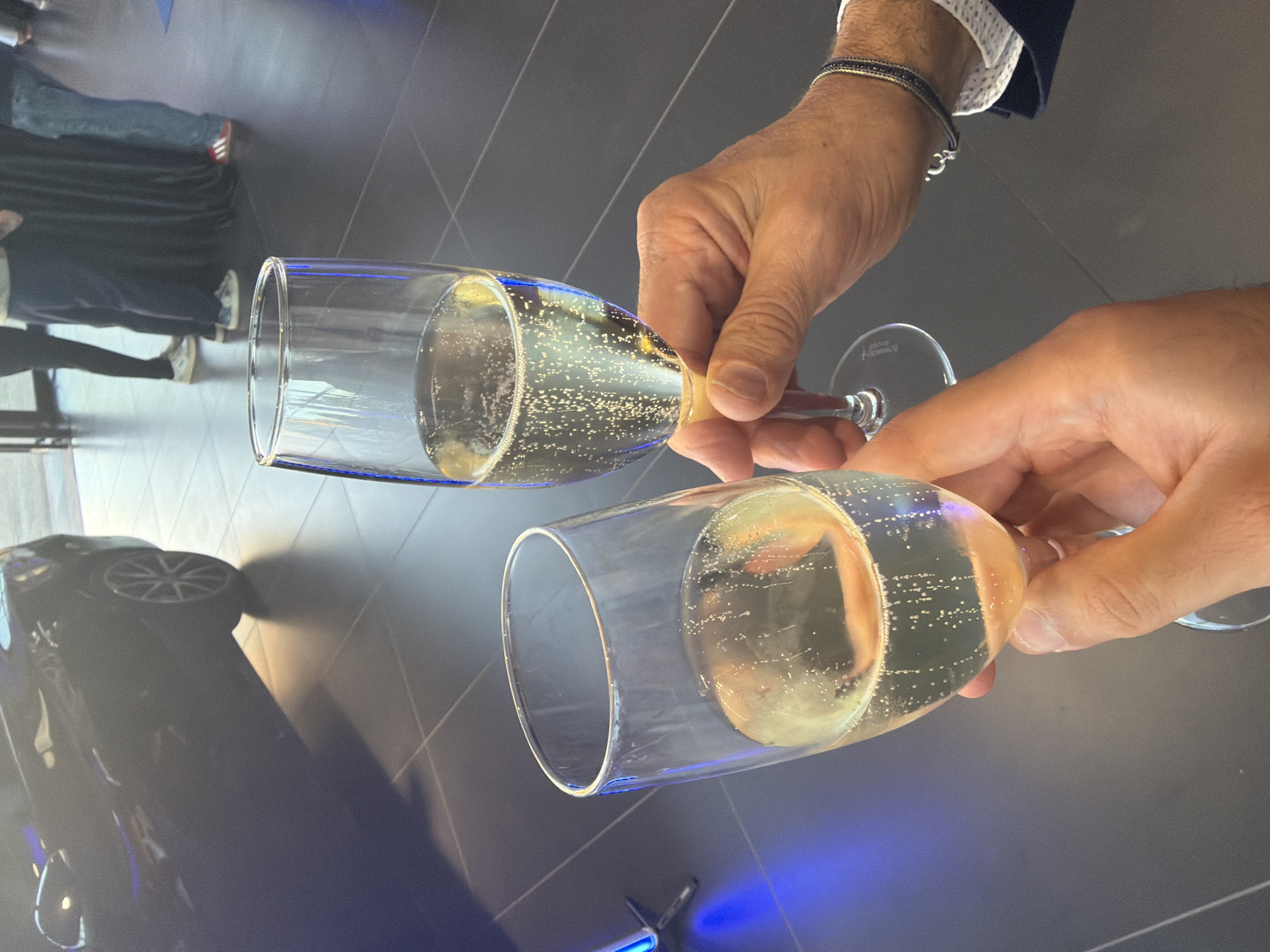 Two champagne flutes filled with a bubbly, pale yellow liquid are being held by two people. The flutes are clear glass with thin stems. The background is dark and appears to be a tiled floor. A dark-colored car is visible in the upper left corner of the image. In the background, there are several people standing, some wearing dark clothing and others in casual attire. The scene suggests a celebration or event, possibly at a car dealership or a formal gathering. The image does not provide any specific visual cues to indicate a location in Parma, Italy.