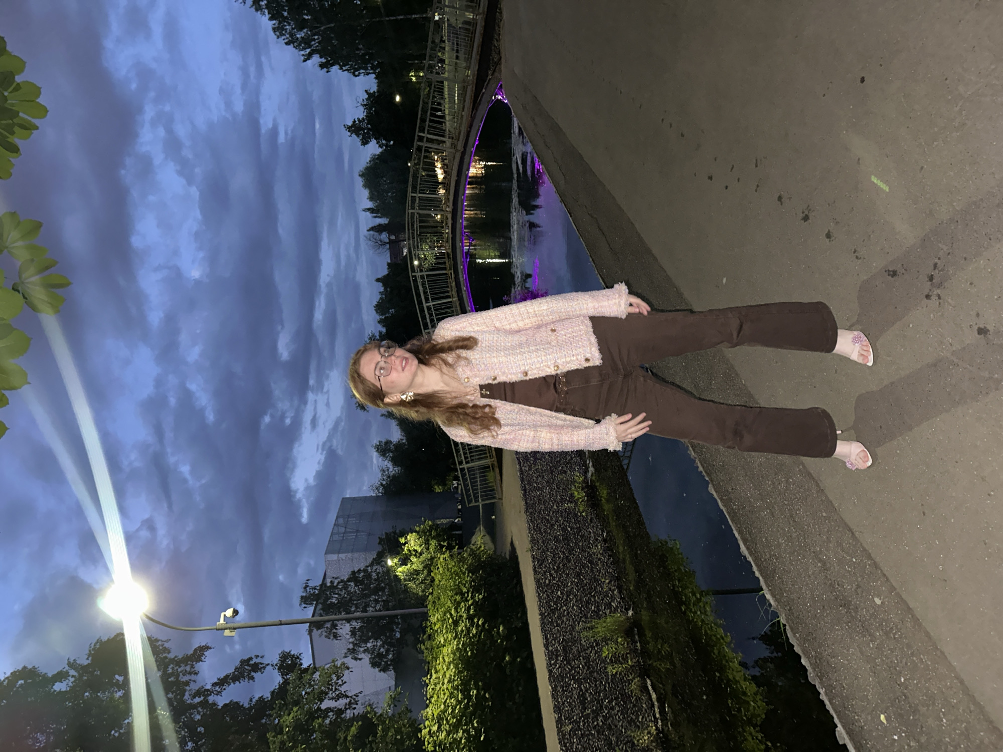 The image depicts a young adult female standing on a paved path next to a narrow canal or stream during twilight or night hours in the city of Kazan, Russia. The sky is overcast with dark blue-grey clouds.
The female subject stands centrally, facing slightly to her right with her gaze directed off-camera. She has long, reddish-brown hair and wears eyeglasses. Her attire consists of a light-colored, possibly pink or cream, textured jacket with gold-colored buttons, worn over a dark brown tank top, and dark brown flared trousers. Her footwear comprises transparent high-heeled sandals with a pink floral embellishment on the front strap. Large, light-colored earrings are visible.
To the left of the subject, a bright street lamp on a pole emits strong light rays; a security camera is mounted below the lamp. Dense green foliage, including bushes and trees, lines the canal. A modern-looking building with a textured, possibly reflective facade is visible in the left background. In the central background, a pedestrian bridge with metal railings arches over the canal, illuminated with purple lighting that reflects on the water's surface. The canal water is dark and reflective. The paved path exhibits minor stains and marks. Shadows are cast by the subject and the streetlight.