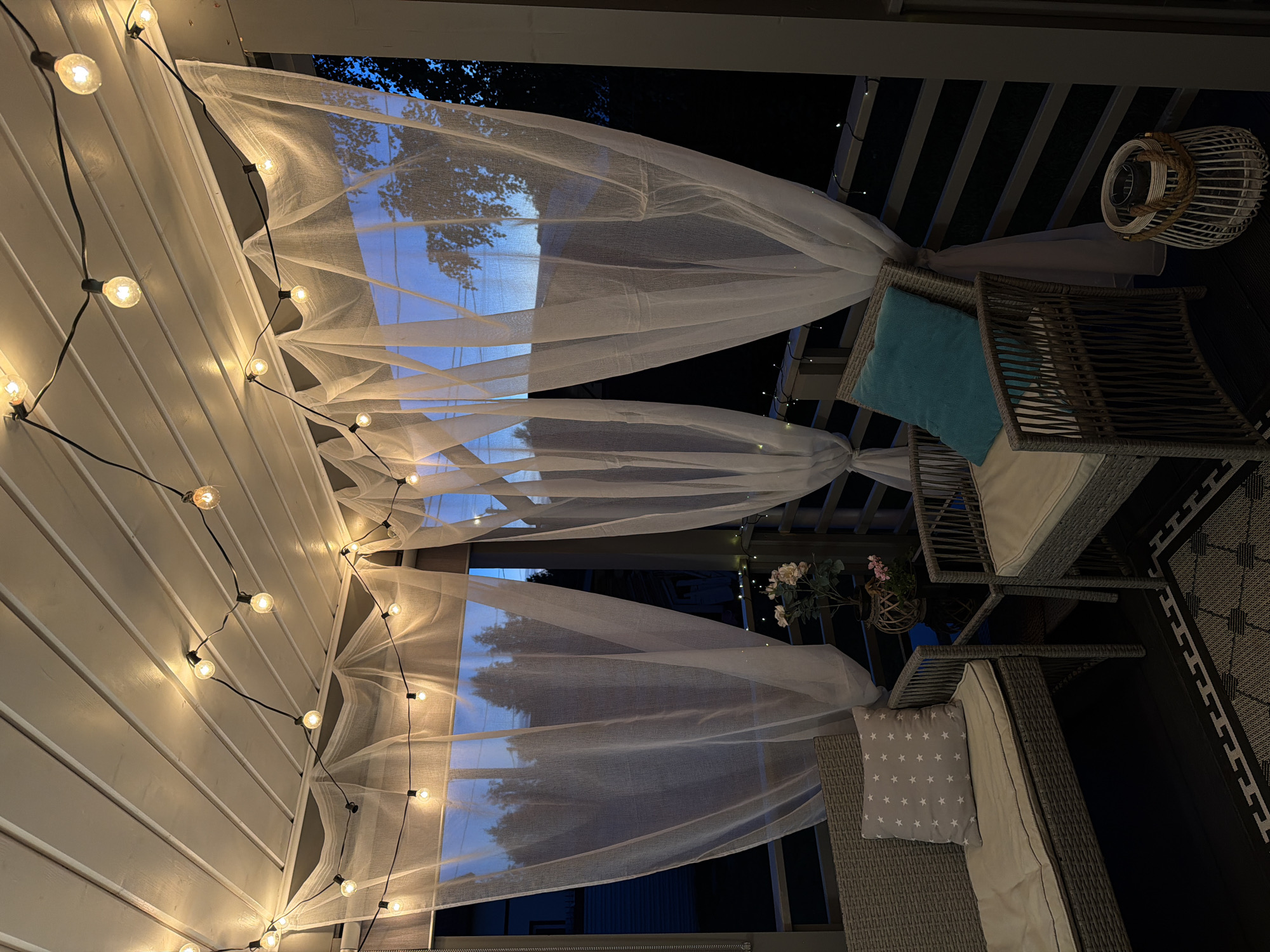 The image depicts a covered outdoor patio or porch area, likely at dusk or night, as evidenced by the warm glow of string lights and the darkening sky visible through sheer white curtains. The patio is furnished with two woven wicker chairs and a matching sofa. The sofa has a light-colored cushion and a grey throw pillow with white star patterns. The chair in the foreground has a light-colored seat cushion and a turquoise accent pillow. A dark rug with a geometric pattern is visible on the floor. A small, dark-colored table sits between the furniture, holding a potted plant with light-colored flowers and another smaller pink flower. A white wicker lantern with rope handles is placed on the floor next to the chair. The ceiling of the patio is paneled with white wood, and the string lights are attached to it, casting a warm illumination. The sheer curtains are draped from the ceiling, creating a soft, ethereal effect and obscuring the view of the exterior, though hints of trees and a building are visible through them. The railing of the patio is dark-colored and appears to have another strand of small fairy lights along it. The overall impression is one of a cozy and inviting outdoor relaxation space, potentially located in the city of Alyoshkino DP, Russia, given the context provided. There are no people visible in the image.