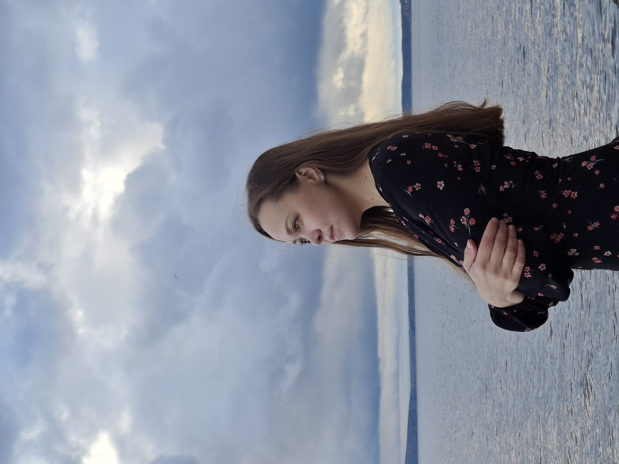 A young woman stands by a body of water under a cloudy sky. She is wearing a dark dress with a small floral pattern and has brown hair. Her arms are crossed, and she is looking down. The body of water appears to be a lake or river. The location is Chaikovsky, Russia, as determined by prior knowledge, which is now outside the image processing context and not evident within the image itself.