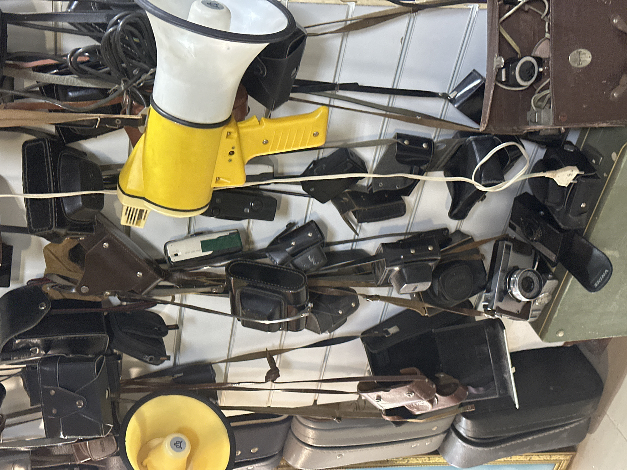 The image shows a collection of vintage camera equipment hanging on a wall. There are numerous black leather camera cases, some with shoulder straps. A yellow and white megaphone is also present. The scene suggests a vintage camera shop or a personal collection. While it may be possible the items are located in Nikolo-Beryozovka, Russia, there is nothing in the photo that confirms its location.