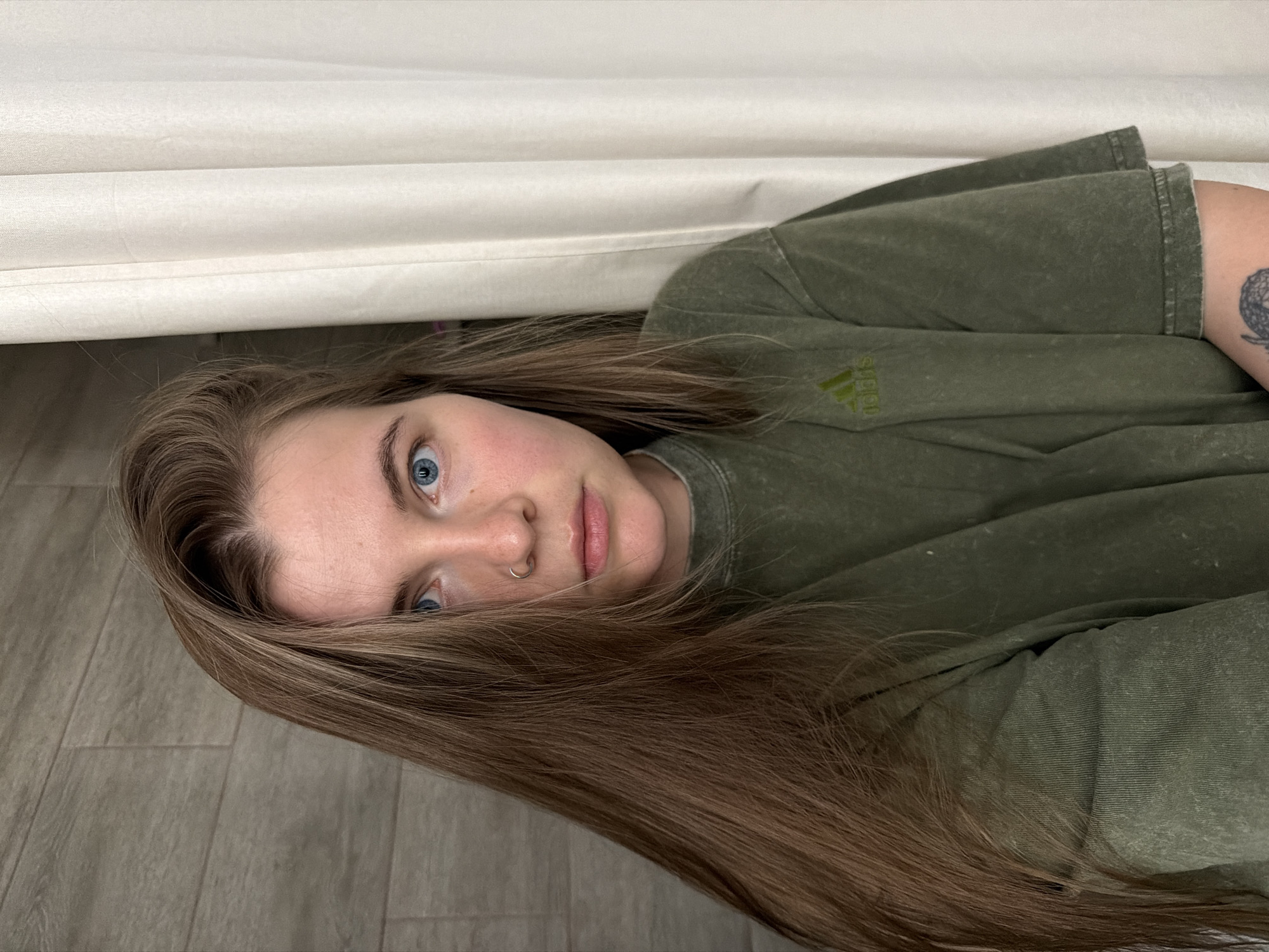 A fair-skinned person with long, brown hair and blue eyes lies on their side on a light gray wood-patterned surface, possibly a floor. A section of the person's face is visible, revealing a nose ring. They are wearing a faded green t-shirt with the word "Adidas" visible on the left breast. Part of the person's left arm is visible and covered with a tattoo of an unknown design. The top of the image is dominated by a beige, fabric-like surface that appears to be draped above the person. The city of Chaikovsky, Russia, cannot be verified based on the visual data.