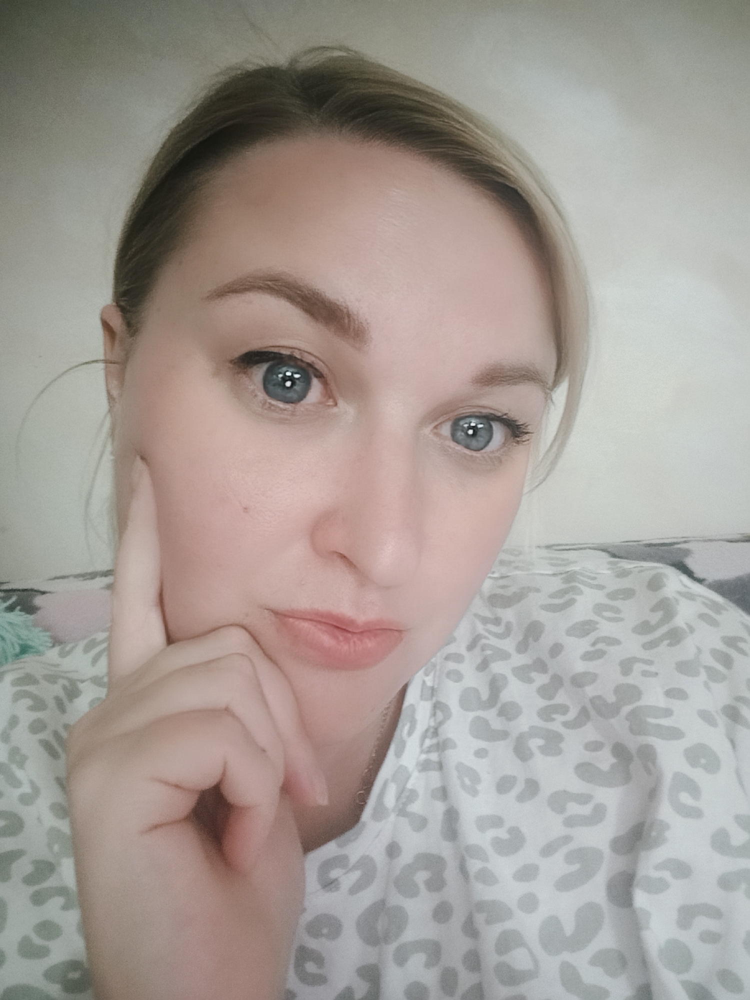 A close-up selfie of a fair-skinned woman with light blonde hair. Her hair is pulled back. She has blue eyes, defined eyebrows, and a neutral expression with slightly pursed lips. Her right hand is raised to her face, with her index finger touching her cheek in a thoughtful gesture. She's wearing a light-colored shirt with a subtle gray leopard print. The background is a blurred, neutral wall and a glimpse of a patterned surface, possibly a blanket or cushion. There is no information about the location of the photo in Chaikovsky, Russia.