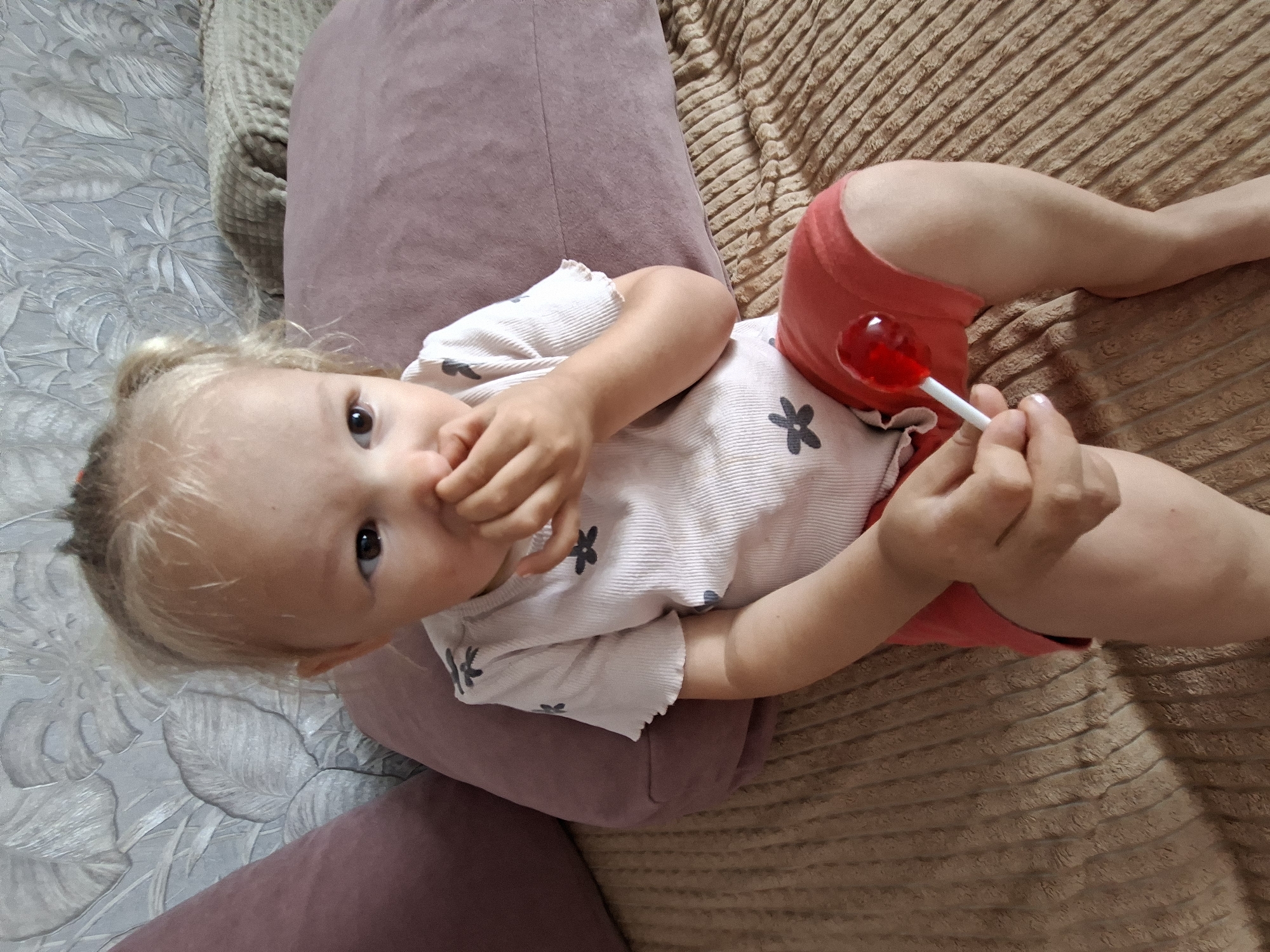 A young child with blonde hair in pigtails sits on a bed, holding a red lollipop. The child is wearing a white t-shirt with a floral pattern and red shorts. Their right hand is near their mouth, and their left hand is holding the lollipop. The background shows a textured wall and pillows. The image is taken from a medium-high angle.