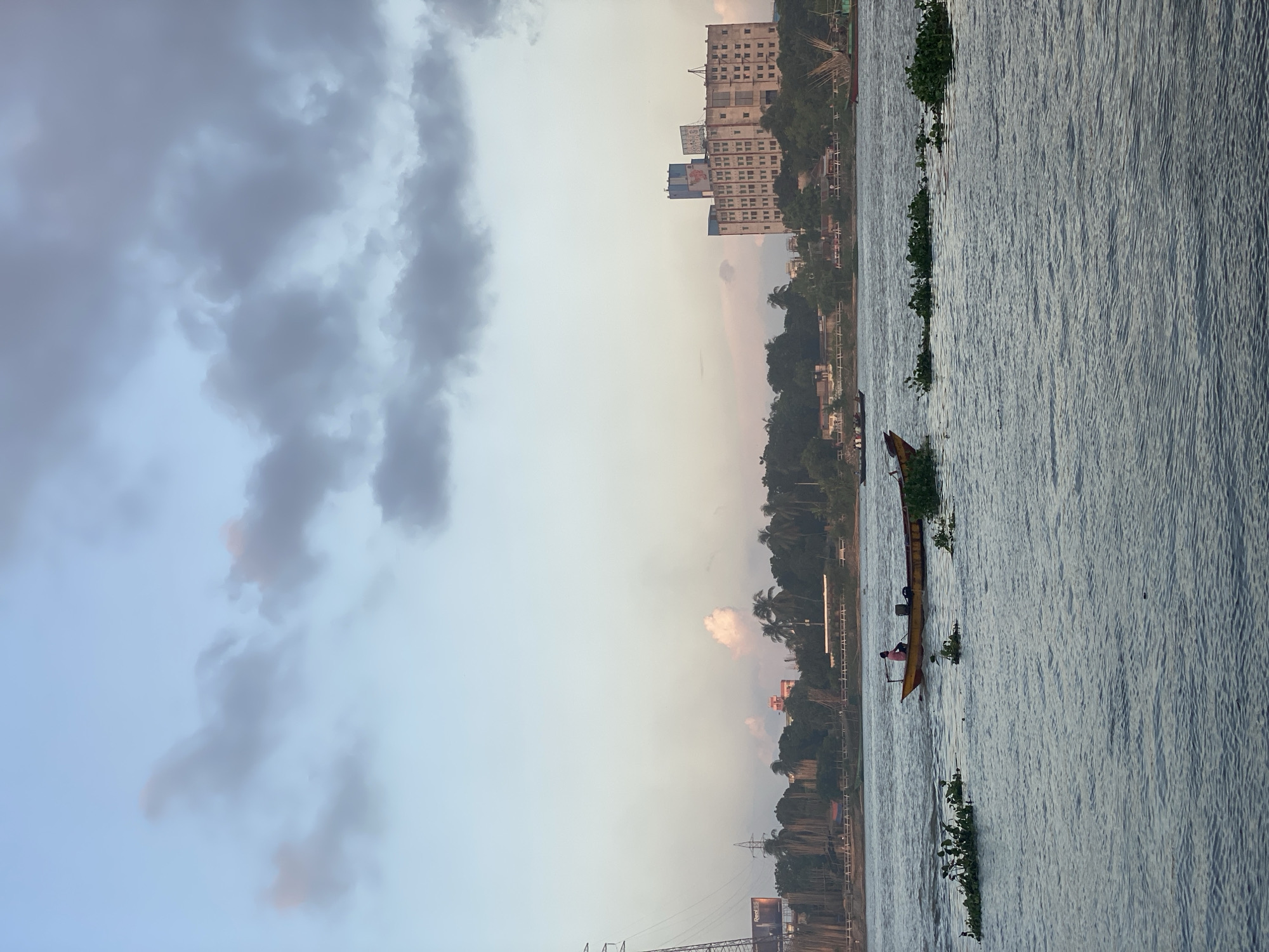 The image depicts a wide, calm river in Dhaka, Bangladesh, during a cloudy twilight. In the foreground, the river's surface is rippled, with clumps of green aquatic vegetation floating on it. A small yellow boat with a person at the stern is moving across the water, leaving a wake. In the mid-ground, the riverbank is lined with lush green trees, including palm trees, and in the background, a large, multi-story beige building with many windows is visible, suggesting an urban or semi-urban setting. A few other boats are also present on the river further in the distance. The sky is filled with a mix of light and dark grey clouds, with hints of pink and orange from the setting sun. Power lines and a large tower are visible on the left side of the image, further indicating an industrial or developed area. The overall impression is one of a peaceful, transitional moment in a riverside cityscape.