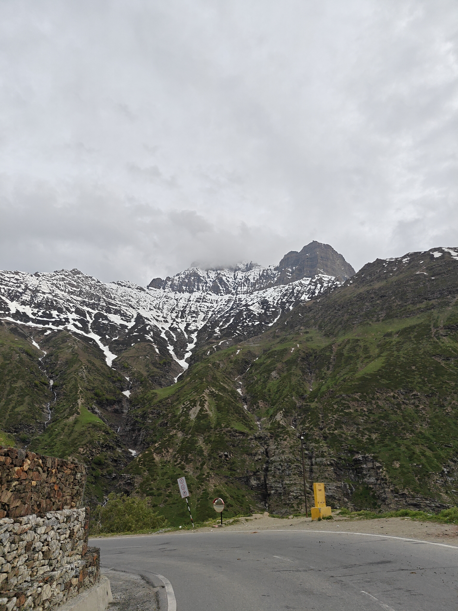 The image shows a mountain range in the vicinity of the city of Shuiling, India. The mountains have a mix of green vegetation and patches of snow. A road runs along the base of the mountains, with a low stone wall to one side. There is a yellow marker and road signs. The sky is overcast.