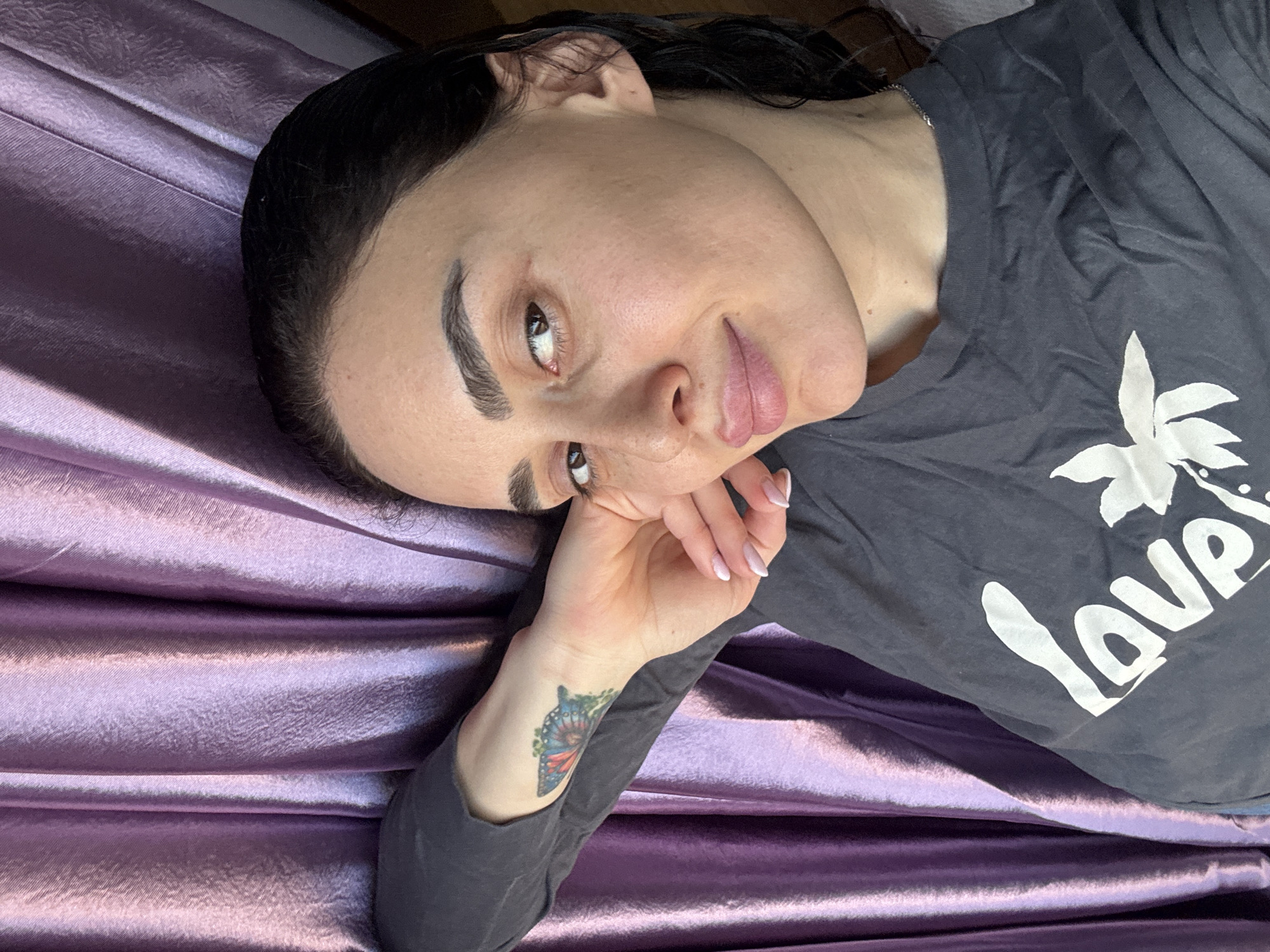 A woman is captured in a medium shot, lying diagonally on a lustrous, draped purple fabric, which appears to be satin. She gazes softly towards the camera with a relaxed expression. Her dark hair is styled back from her face, which appears natural with prominent eyebrows. She wears a dark grey t-shirt featuring a white graphic print that includes palm fronds and the partially visible word "Love."
Her left hand, adorned with an ombre pink and white French manicure, gently touches her chin or cheek. A vibrant butterfly tattoo, showcasing shades of blue, orange, and green, is visible on her left wrist. The purple fabric beneath her is folded and creased, creating variations in light and shadow. The setting appears to be indoors, possibly on a bed or sofa, conveying a calm and intimate atmosphere. The location context for this image is Tbilisi, Georgia.