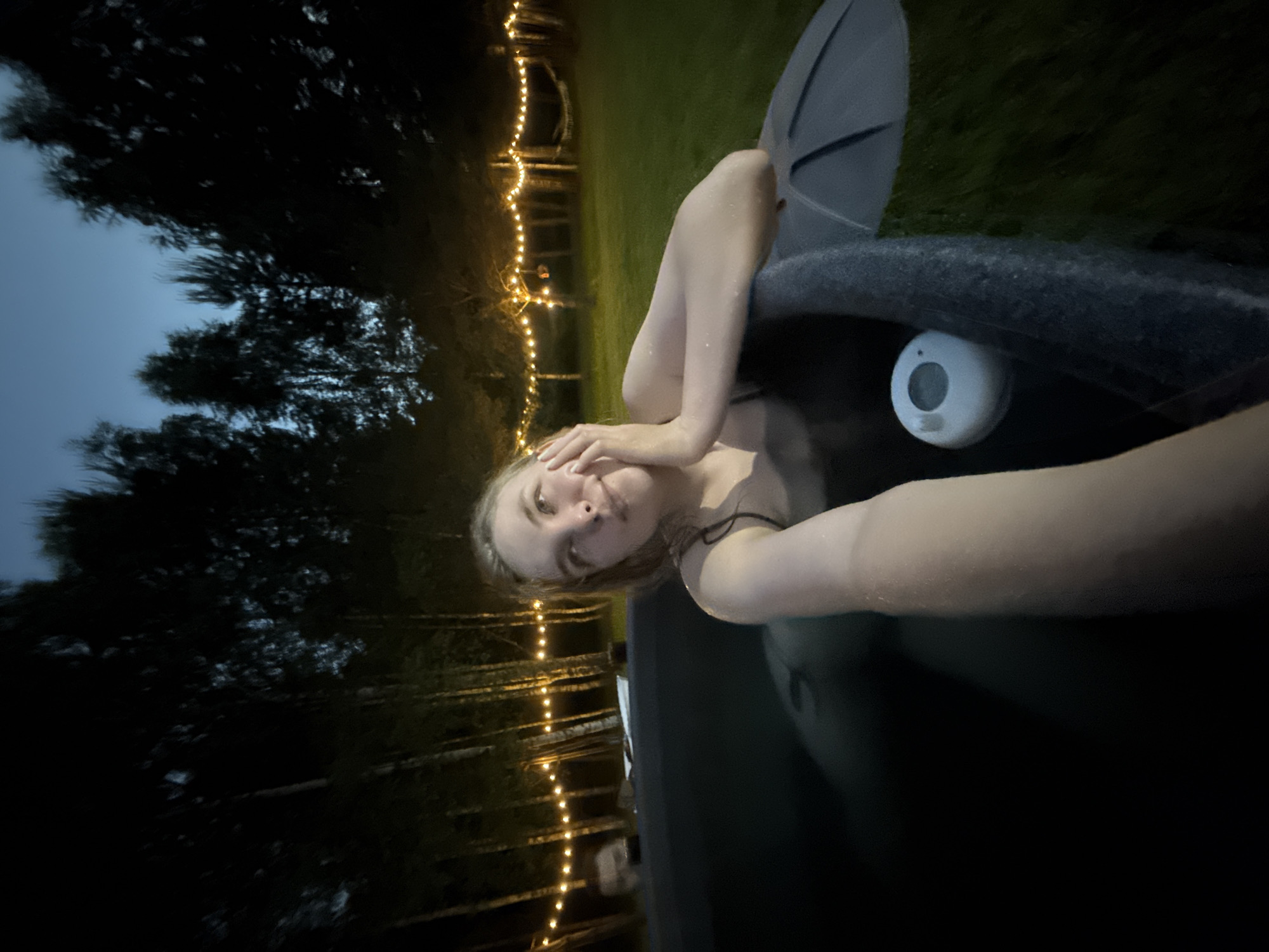 An outdoor nighttime scene depicts a young woman in an inflatable hot tub. The woman, with light-colored, damp hair and wearing a dark-colored top, is positioned in the tub with her right arm resting on the rim and her left hand touching her left cheek. Her gaze is directed upwards and slightly to the right. A white, circular floating device with a display is visible in the water to her right. The hot tub is dark gray. To the immediate right, a dark gray section of what appears to be a hot tub cover or an adjacent umbrella is visible.
The background consists of a grassy area, illuminated by a string of warm yellow lights strung between numerous trees with slender, light-colored trunks, indicative of birches. The sky above is dark blue, suggesting late dusk or night. A faint, partially submerged head of another individual is discernible in the water behind the primary subject, towards the left of the frame. Further into the background, obscured by trees, are indistinct shapes and possibly a hammock or other outdoor furniture. The scene is located in Копнино, Russia.