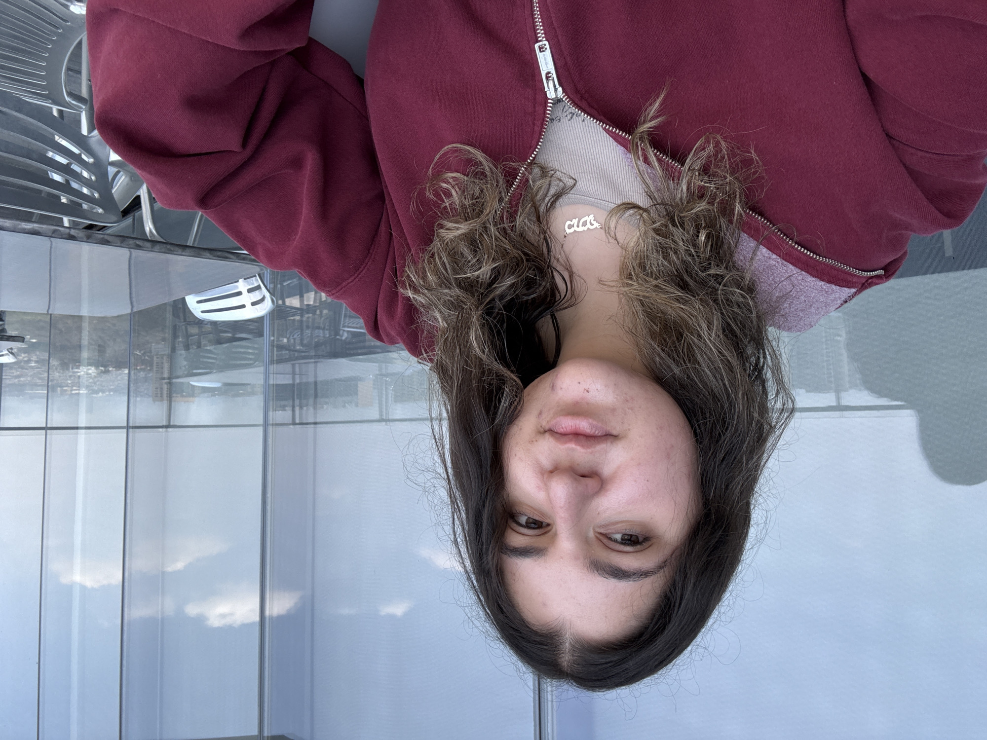 A woman with brown hair is wearing a red zip-up sweatshirt and a necklace that says "Chloe." She is inside a building with large windows offering a view of Mexico City. The windows reflect the sky and clouds. There is a table and chair in the foreground.