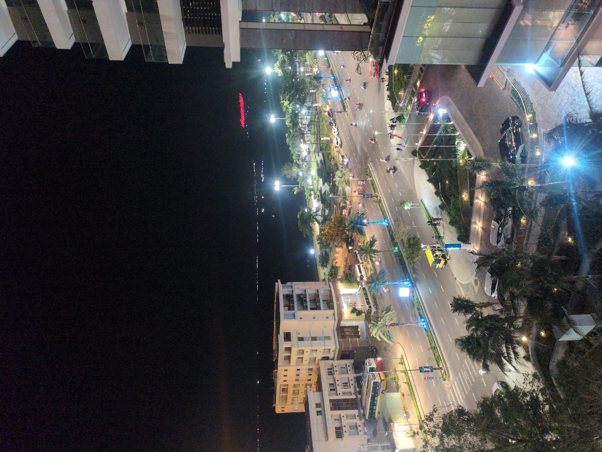 An elevated nighttime photograph depicts a bustling urban street scene in South Quy Nhon Ward, Vietnam. The image captures a wide, multi-lane asphalt road, divided by a landscaped median strip containing numerous palm trees, running horizontally across the mid-ground. This road parallels a calm body of water, likely the ocean or a bay, visible on the left, reflecting scattered lights from distant landmasses and vessels.
The environment is heavily illuminated by artificial light sources, including numerous tall streetlights casting bright white light, vehicle headlamps and taillights, and exterior lighting from buildings. The palm trees along the median and sidewalks are adorned with decorative blue lighting.
Traffic on the main road is active, consisting primarily of numerous motorcycles and scooters, with several passenger cars and at least one long, red and yellow public bus visible on the left side of the road. A yellow four-wheeled vehicle, possibly a taxi, is stopped near the curb on the right side. White lane markings and crosswalks are discernible on the asphalt. Several individuals are operating motorcycles, and a few pedestrians are observed on the sidewalks and near crosswalk areas, indicating typical urban nighttime activity.
To the immediate right in the foreground, a modern multi-story building with a glass facade and white balconies is partially visible, with an illuminated driveway below where a dark car is entering. To the left of the main road, closer to the water, stand multiple other multi-story buildings, predominantly white or light-colored, featuring balconies. In the far background, across the bay, a large red illuminated sign, partially legible as "FLC Luxury Resort," is visible against the dark horizon.