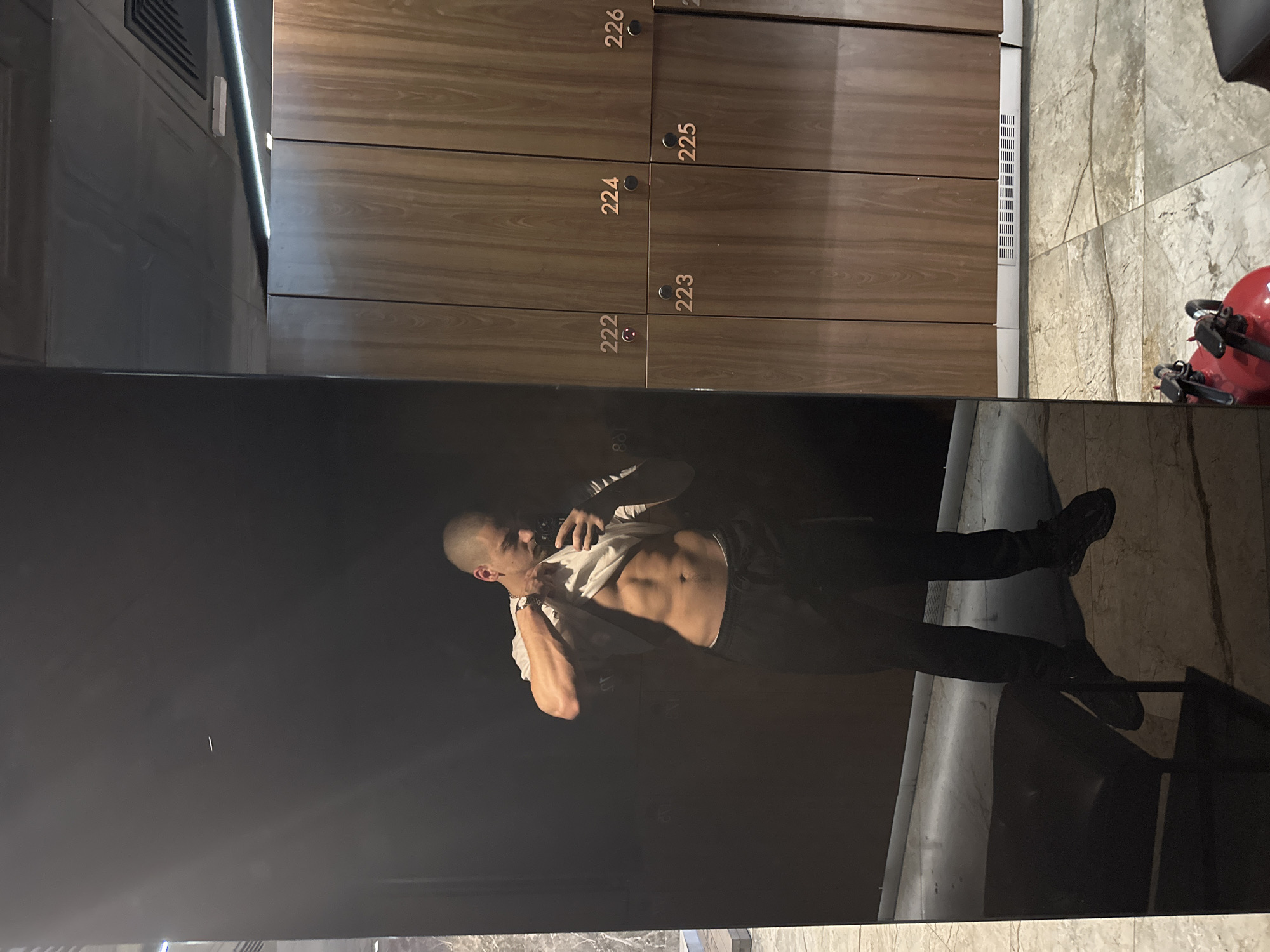 A man takes a selfie in front of a black mirror in what appears to be a locker room. The lockers in the background are numbered 222 through 226. A fire extinguisher is also visible.