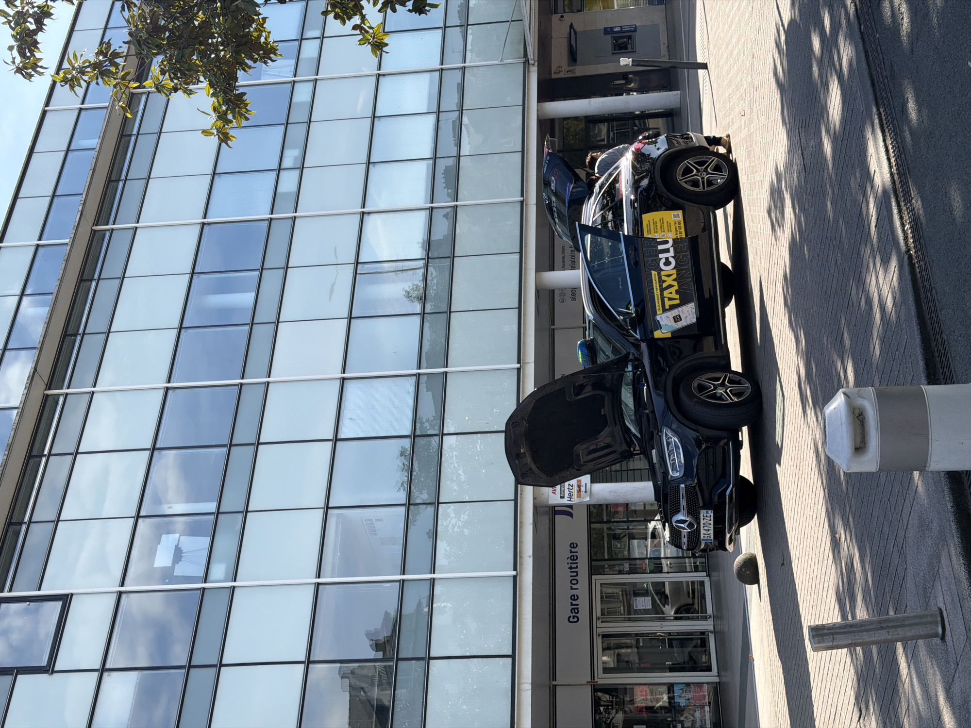 The image depicts a black Mercedes SUV with its hood and doors open parked on a sidewalk adjacent to a building. The building's facade is predominantly glass with square windows. The words "Gare routière" are visible above the entrance, along with signs for "Europcar," "Hertz," and "Alep." The SUV has a "TAXICLUB ANGERS" advertisement on the side, with a telephone number. A person appears to be inside the vehicle. The license plate reads "GX-470-ZE." The setting suggests the location is near the Angers bus station in France. Shadows cast by trees are visible on the sidewalk. A gray cylindrical object is located near the edge of the sidewalk.