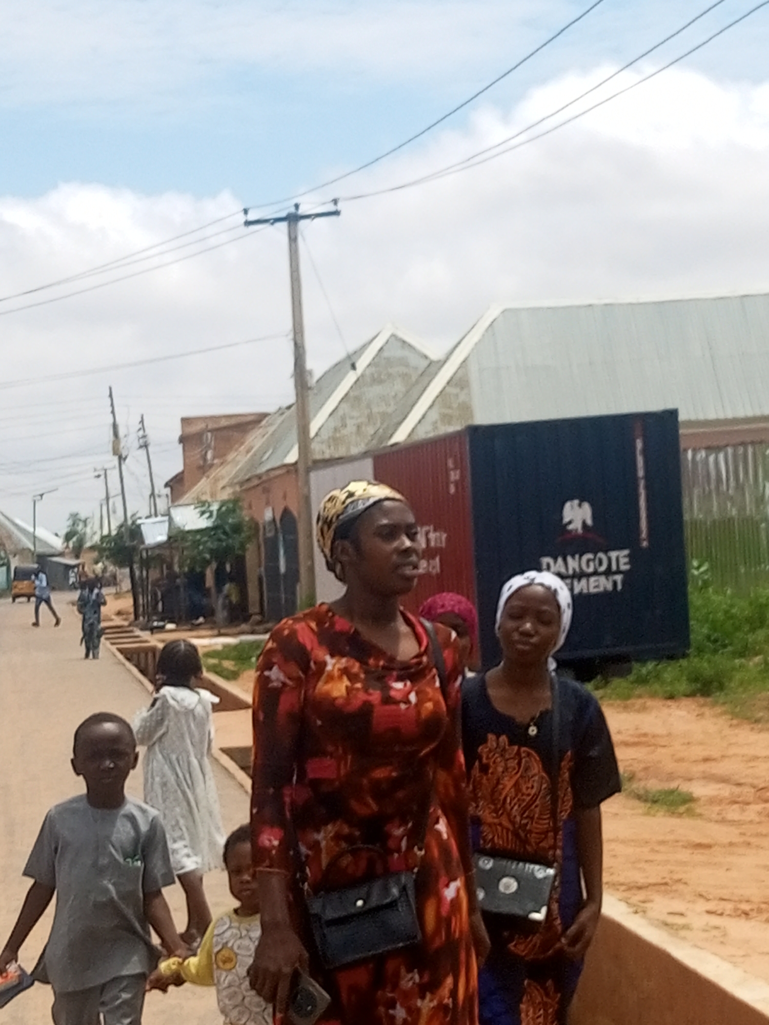 The image captures a street scene in Tempure, Nigeria, under a bright, overcast sky. In the foreground, a woman in a vibrant, patterned dress is positioned to the right, her gaze directed slightly downwards. To her left, a young boy in a grey tunic and trousers walks forward, holding a small item in his left hand. Another young child, wearing a yellow and white patterned shirt, stands next to the boy, holding his hand. Further back, a person in a white garment walks away from the camera, and other figures are visible on the street in the distance.
The environment consists of a dirt road lined with buildings and structures. Visible in the background are several buildings with corrugated metal roofs. A large, dark blue shipping container with white lettering, including the word "DANGOTE," is prominent on the right side of the image. Power lines and utility poles are also present, indicating urban infrastructure. The overall impression is that of a typical day in a Nigerian town.