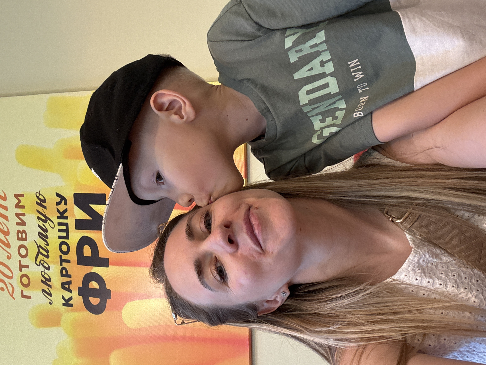 A woman with long blonde hair and fair skin is pictured in a close-up shot with a young boy wearing a black cap and a green shirt with the words "LEGENDARY BORN TO WIN." The boy appears to be kissing the woman on the forehead. Behind them, there is a sign in Russian with an image of french fries, which says, "20 лет готовим любимую картошку фри." This translates to "20 years of making our favorite french fries." Considering this sign in Russian, the location is possibly in Chaikovsky, Russia, although this cannot be confirmed.