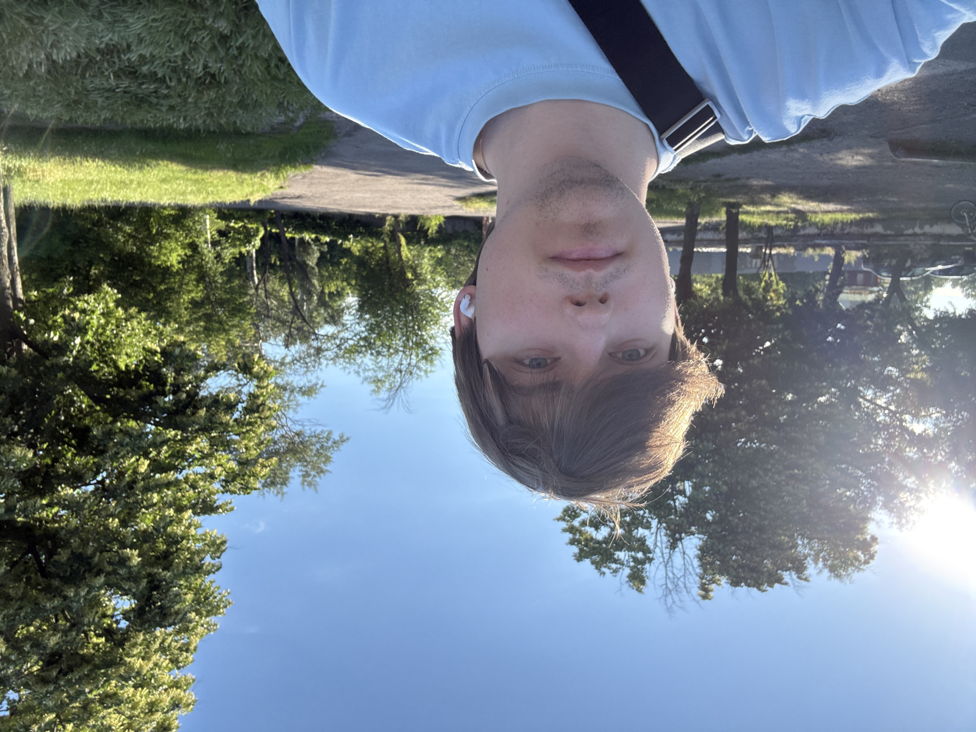 A young man with light brown hair and blue eyes is taking a selfie outdoors. He is wearing a light blue t-shirt and a black strap is visible across his shoulder, suggesting a bag. He has a white wireless earbud in his right ear. The background shows a clear blue sky with the sun shining brightly from the left, casting lens flare. There are several trees with green foliage, and a paved path is visible in the middle ground. In the far background, cars are parked and some buildings can be seen. The environment appears to be a park or an open area within a city, possibly in Dnipro, Ukraine, given the vegetation and typical urban setting. The lighting suggests it is daytime, likely late afternoon or early morning due to the angle of the sun.