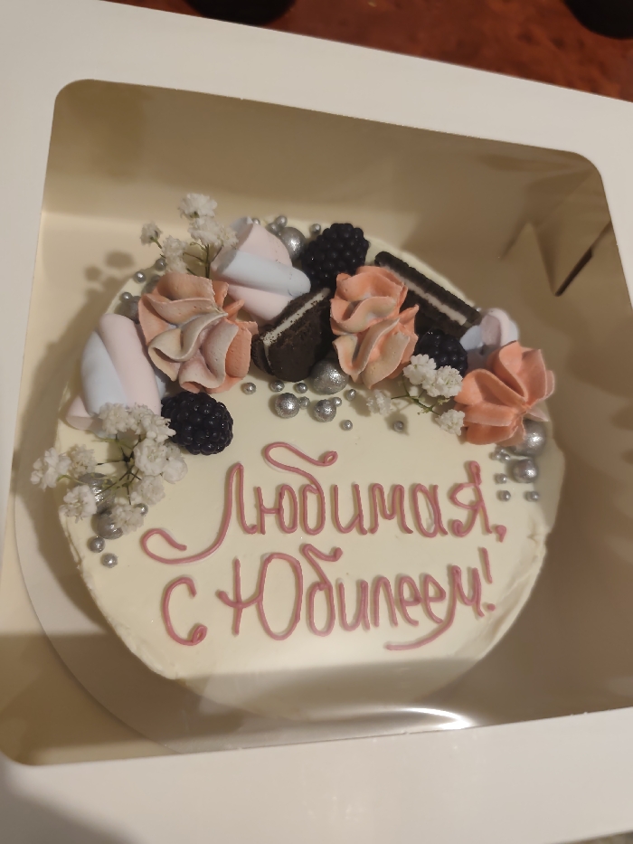 A round celebratory cake, frosted in white, is depicted inside a white cardboard bakery box with a clear plastic top section. The cake's surface is decorated with clusters of pink and white swirled buttercream, whole blackberries, fragments of chocolate sandwich cookies, and scattered silver edible pearl sprinkles. Small clusters of white baby's breath flowers are also arranged on top. Written in pink script across the cake is the Cyrillic phrase "Любимая, с Юбилеем!" (Beloved, Happy Anniversary/Jubilee!). The cake box rests on a dark, unidentifiable surface, possibly a wooden tabletop. This scene is located in Chaikovsky, Russia.