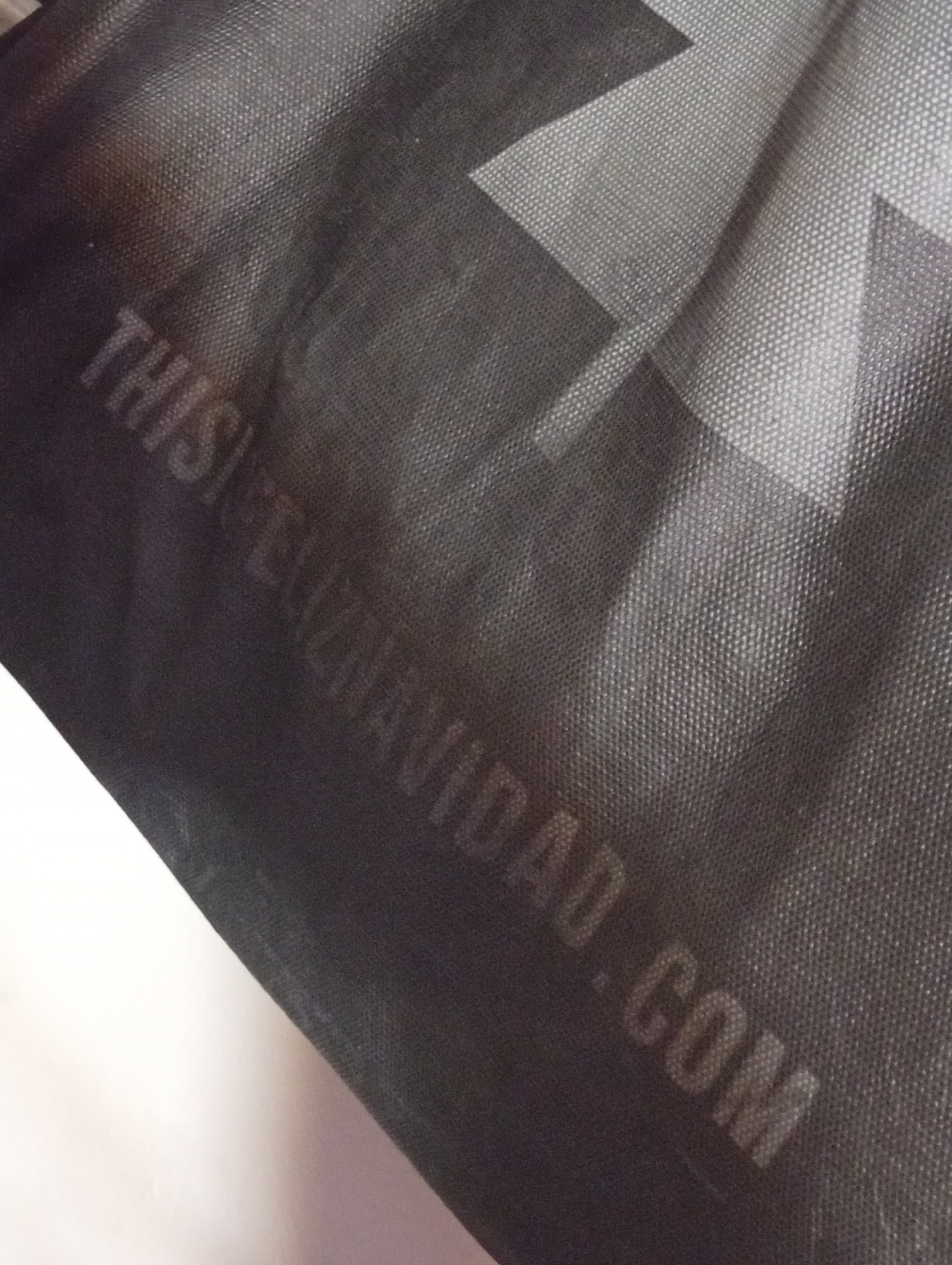 The image shows a close-up of a dark grey or black tote bag made of woven material. In the background, there is a portion of a white surface. The text "THISI FELIZNAVIDAD.COM" is faintly printed on the bag. There is also a larger, lighter grey chevron or cross-shaped pattern partially visible on the bag. The image provides no information indicating a location in the city of Buenos Aires, Argentina.
