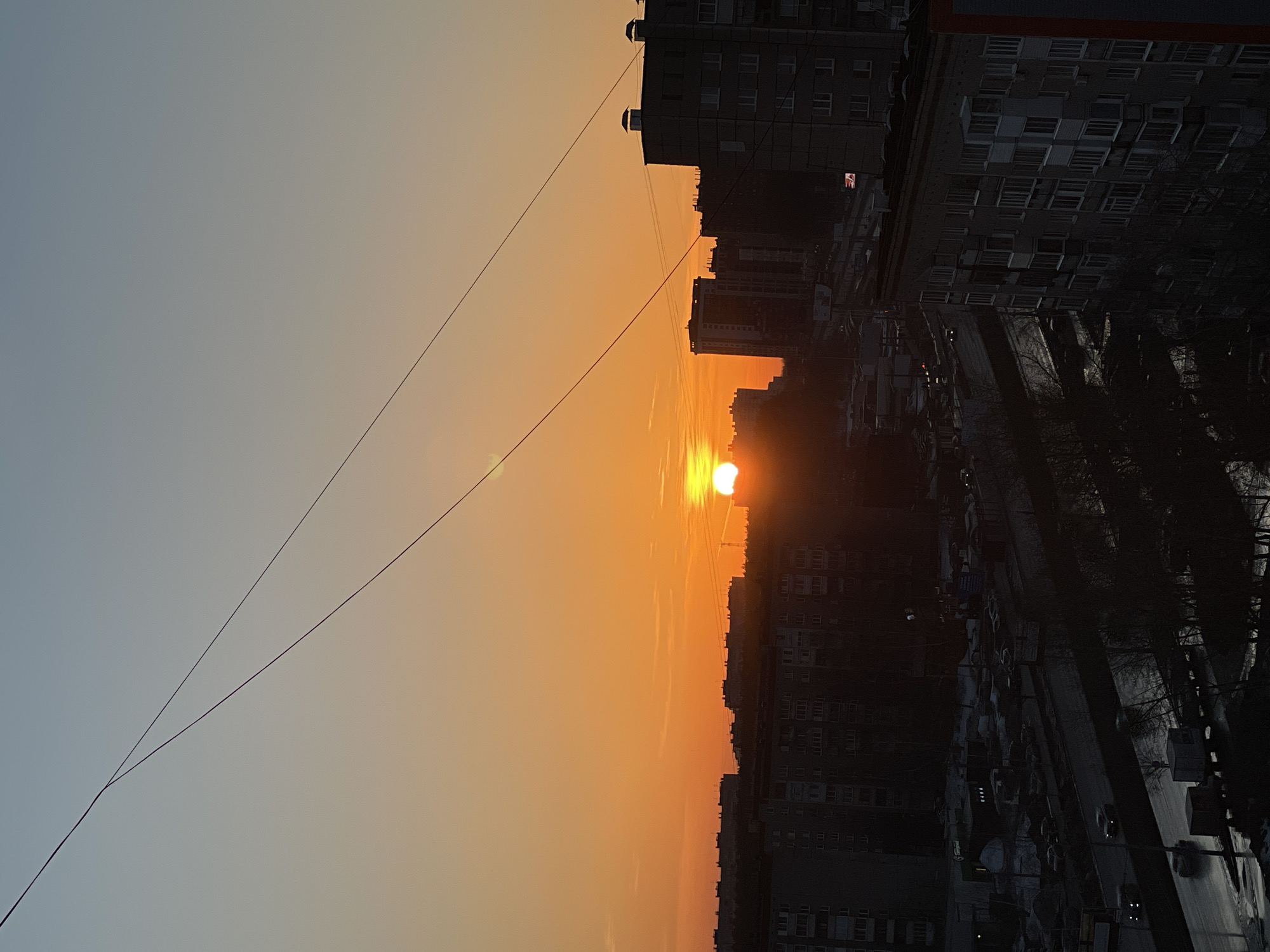 The image shows a sunset in Izhevsk, Russia. The sun is setting over the city skyline, silhouetting buildings. In the bottom right portion of the image, a road and sidewalk are visible, with cars and trees lining the side. Two power lines stretch across the horizon, intersecting slightly to the left of the image's center. The sky transitions from light blue in the upper-left to bright orange around the setting sun.