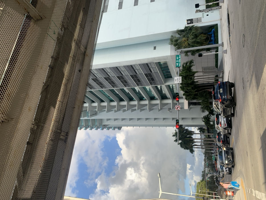 The image presents an urban street view captured from beneath an elevated structure featuring a wire mesh ceiling, likely an overpass or a parking garage entrance. The scene is located at the intersection of NE 2nd Avenue and Abel Holtz Boulevard in Miami, United States of America, as indicated by a green street sign.
In the foreground, a multi-lane road is visible, where approximately seven vehicles are stopped in traffic, predominantly dark-colored SUVs and sedans. The traffic light at the intersection displays a solid red signal for vehicles approaching the intersection. A white "NO TURN ON RED" sign is mounted below the traffic light, alongside a "no left turn" symbol. To the far left, a light-skinned pedestrian carrying multiple shopping bags in their left hand walks on the sidewalk, moving towards the background.
The right side of the frame is dominated by a modern, light blue and grey multi-story high-rise building with numerous balconies featuring glass railings and large, rectangular windows. The building exhibits a stepped or staggered architectural design on its facade. Palm trees are strategically planted along the street and around the base of the high-rise building. A blue, abstract sculptural object is visible on a landscaped patch near the sidewalk at the base of the tall building. The sky above is partially cloudy with patches of clear blue. Street lighting fixtures are visible on both sides of the road.