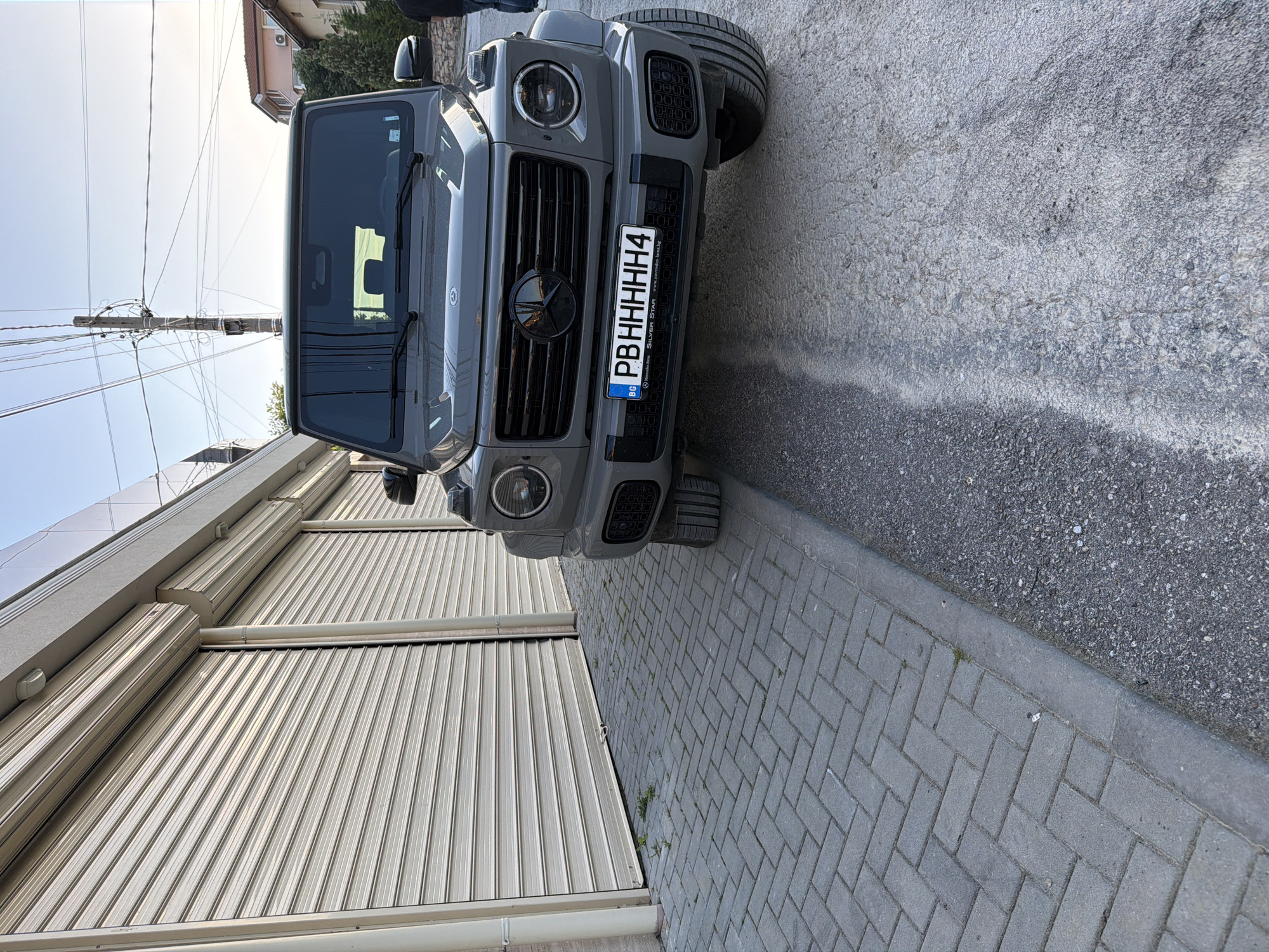 The image depicts a grey Mercedes-Benz G-Class parked on a street in Asenovgrad, Bulgaria. The car is positioned at an angle, facing towards a closed metal garage door. The front of the vehicle is visible, including its grille, headlights, and license plate, which reads "PBHHHHH4" with the Bulgarian flag. The background includes a grey brick sidewalk, asphalt pavement, and a building with a beige facade. A utility pole with multiple wires is present in the upper left corner of the image.