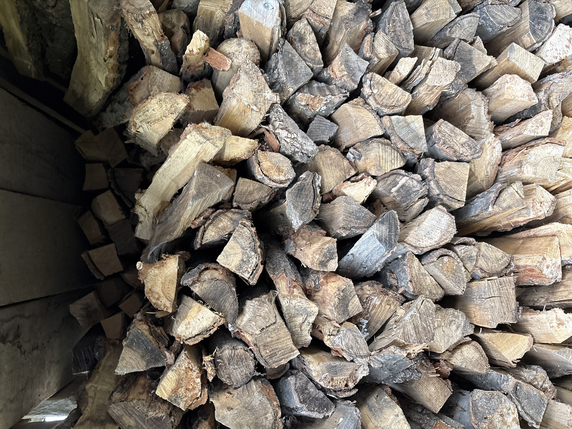 The image is a close-up, vertical shot of a large stack of cut firewood. The wood is primarily composed of logs split into various sizes, with some larger, rougher pieces visible. The bark is still attached to many of the pieces, and the wood shows evidence of age and weathering, with variations in color from light brown to dark gray. The grain of the wood is clearly visible, with rings and cracks indicating the material's organic nature. The firewood is stacked densely, filling the frame, with the top of the stack meeting a dark, flat surface, possibly the underside of a roof or shelter. The lighting is somewhat dim, casting shadows and highlighting the textures of the wood. There are no people or animals present in the image. The scene appears to be an outdoor storage area for firewood. The location is specified as Siniy Gay, Russia.