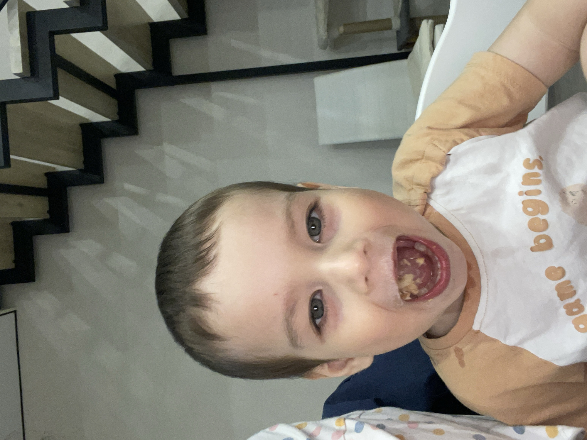 A close-up, portrait-oriented photograph features a young child, likely a toddler, with short brown hair and striking blue-grey eyes. The child looks directly at the camera with a wide-open mouth, revealing partially chewed food inside, possibly a soft grain or fruit mix. There is a small amount of food residue or drool on the child's chin and the upper part of their shirt, suggesting an active eating moment. The child's expression conveys joy or excitement.
The child is wearing a t-shirt with a white torso and orange raglan sleeves and collar. The phrase "game begins" is printed in orange capital letters on the white section of the shirt.
In the background to the child's left, a modern staircase is visible, characterized by dark, angular structural elements that appear to be metal or dark wood, contrasting with lighter-colored treads. Shadows from the staircase are cast on a light-colored wall. To the child's right, a part of a white or light-colored furniture piece, possibly a chair or high chair, is discernible. In the immediate foreground, a blanket or sheet with a colorful, abstract pattern is partially visible.
The scene captures a candid indoor moment, likely during mealtime, in Izhevsk, Russia.