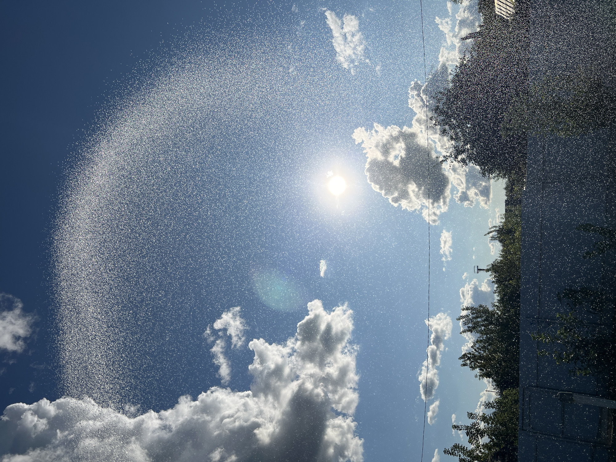 The image depicts a bright, sunny day with a clear blue sky and scattered white clouds. Sunlight streams through the sky, creating a bright glare. In the foreground, a fine mist of water particles is visible, suspended in the air and catching the light, forming a semi-circular arc across the upper portion of the frame. This water mist is also visible throughout the lower part of the image, obscuring a fence and trees. The background shows a line of green trees and a dark fence running horizontally across the lower third of the image. There are no people visible. The scene suggests a residential area, possibly a garden or backyard, in Salavat, Russia.