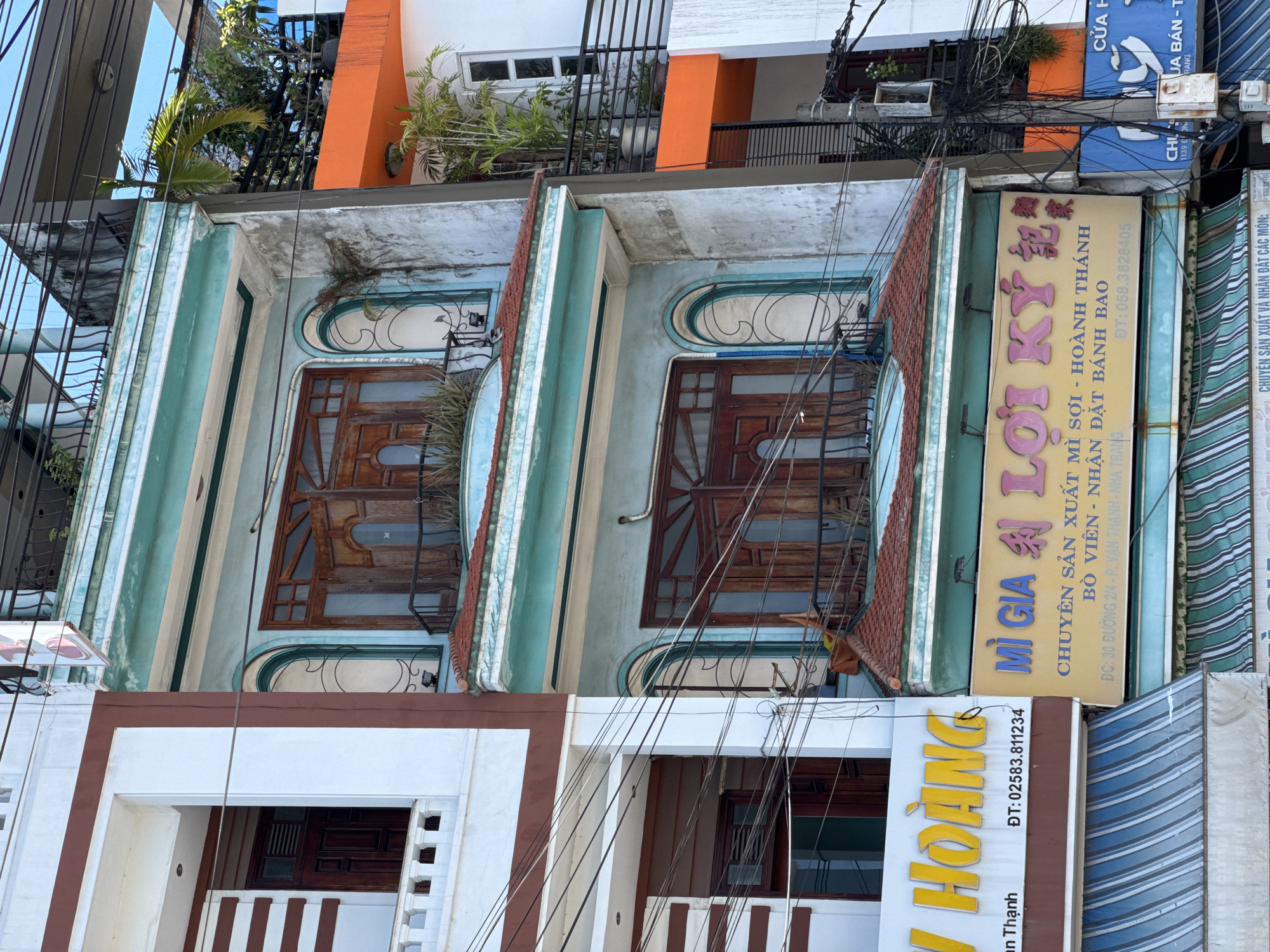 The image shows a narrow, multi-story building facade in Nha Trang Ward, Vietnam. The building has a light blue and white exterior with ornate window frames and wooden doors. There are balconies with metal railings and tiled roofs. Several signs are visible, including one in yellow with Vietnamese text that reads "MÌ GIA LỢI KÝ" and another in blue with Japanese characters and Vietnamese text. The building is surrounded by electrical wires, typical of urban Vietnamese street scenes. The street level appears to have awnings for shops. The overall impression is of a densely built urban environment with a mix of traditional and modern architectural elements.