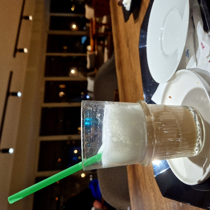 A glass sits on a wooden table, filled with a milky, foamy beverage and a green straw. Beside the glass, stacked white plates rest on a black serving tray. The background shows a blurry interior of a building or cafe, with a series of lights along a wall. The overall image suggests an indoor setting, possibly a restaurant or coffee shop.
