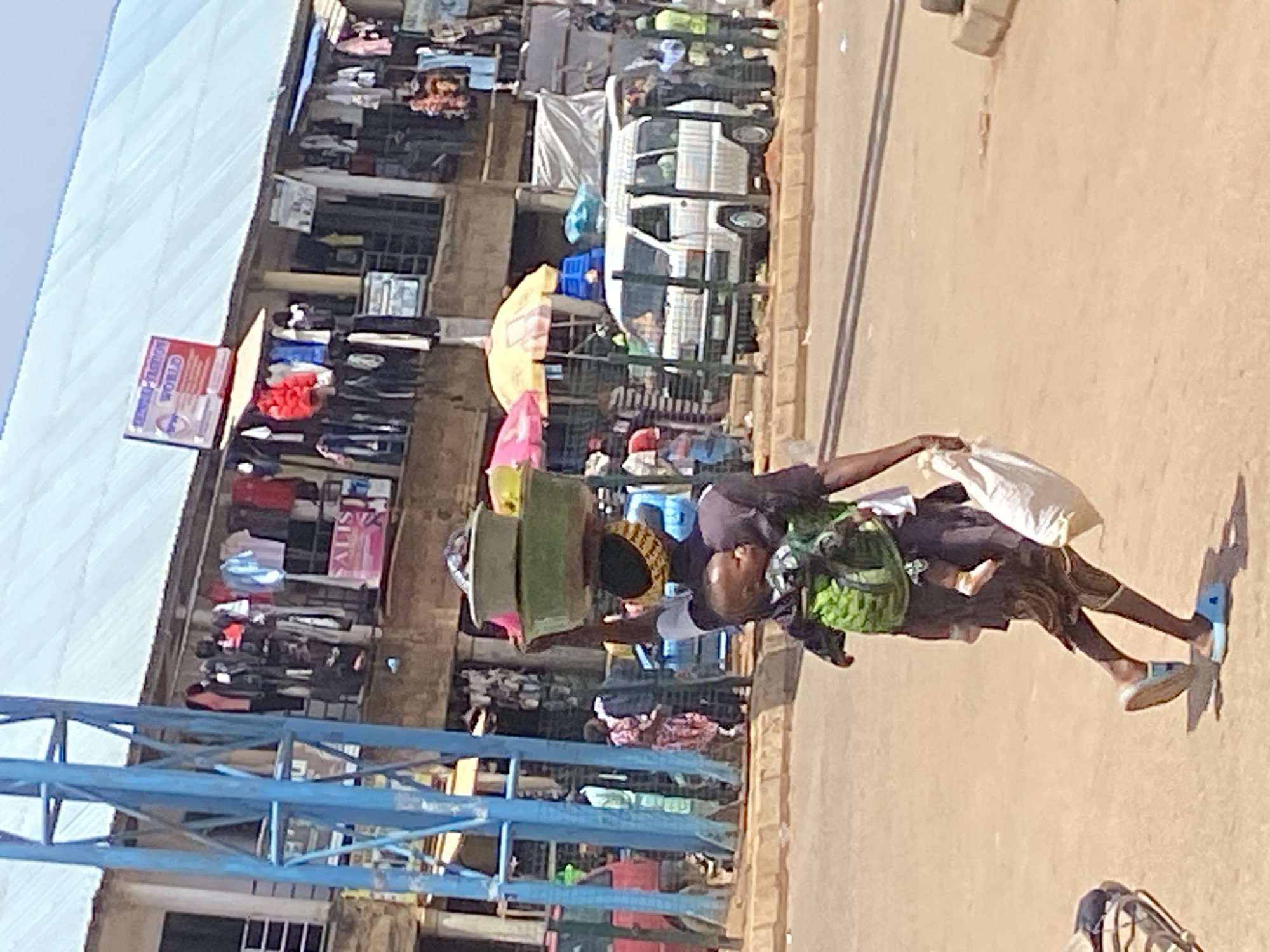 A man walks down a sidewalk in Bukuru, Nigeria. He carries several green rectangular objects on his head. He wears a green backpack and carries a white bag. Buildings and shops line the street. A white van is parked on the street. A blue metal structure is visible in the foreground.