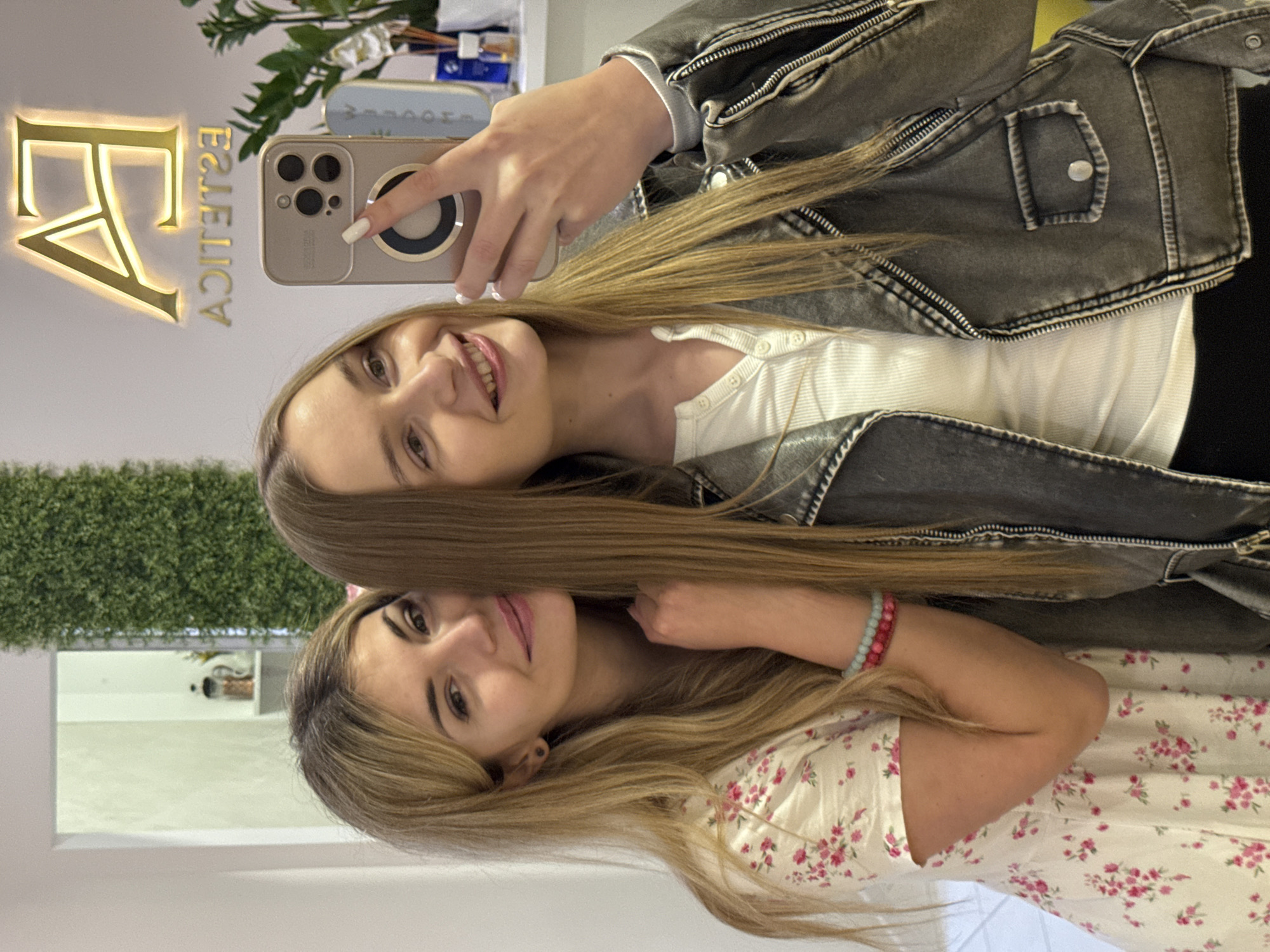Two young women pose for a selfie in what appears to be the Estetica salon, based on the illuminated "EA ESTETICA" sign in the background. The upper woman is holding a phone, likely taking the photo, and is wearing a white ribbed top and a grey denim jacket. The lower woman is wearing a white floral dress and a beaded bracelet. Their blonde hair blends in the middle of the frame. Behind them, a green wall and some decorative plants are visible. The scene suggests a casual, friendly atmosphere inside the salon. The city is implied to be Budva, Montenegro, due to the salon's possible location.