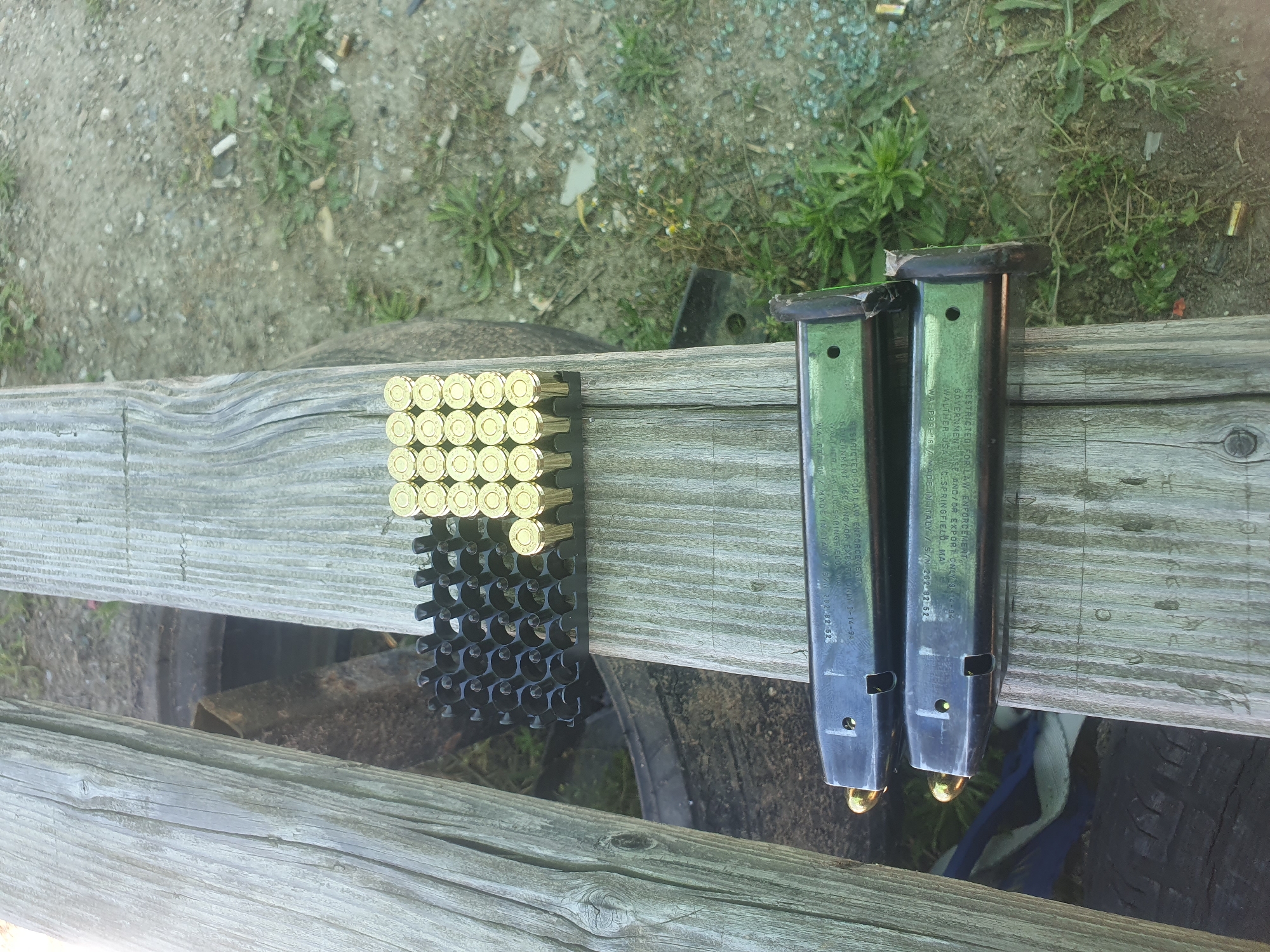 The image depicts ammunition and two magazines on a weathered wooden surface. There is a plastic holder with approximately 50 rounds of ammunition, and two black magazines, each loaded with at least one round. The background shows grass and gravel. There is no indication of the location being Войводино, Bulgaria, or the presence of people.