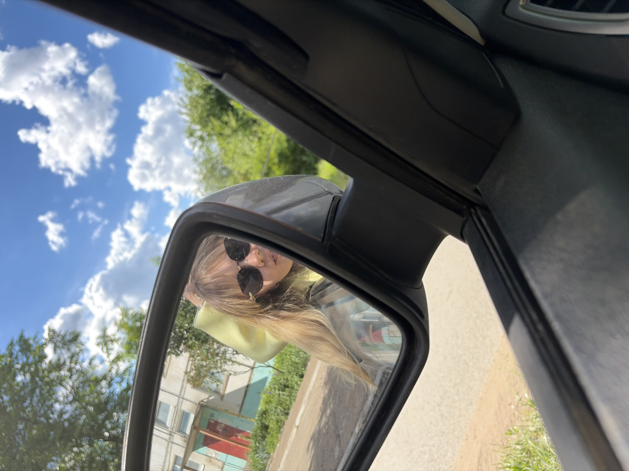 The image shows a view through the side mirror of a car, seemingly parked or stationary. The mirror reflects a young woman wearing sunglasses and a yellow shirt, who appears to be inside the car. In the background visible in the mirror, there's a building, likely residential, along with greenery suggesting a street or park setting. The sky above is blue with scattered white clouds. The location is identified as Chaikovsky, Russia, based on additional context. The car's interior framework surrounds the mirror, providing a partial view of the vehicle's interior.