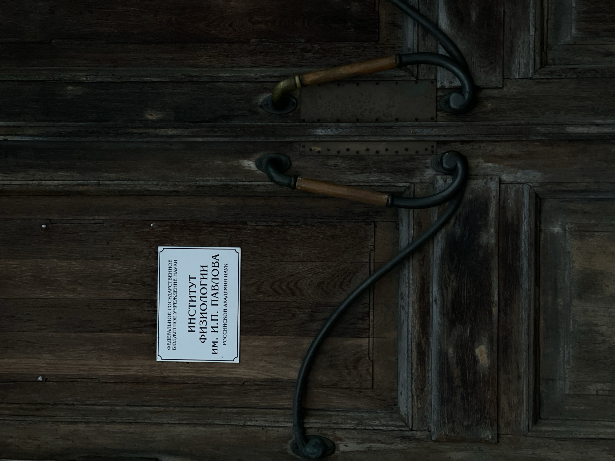 The image shows a close-up of a weathered, dark wooden double door, likely the entrance to a building. A white sign with Russian text is affixed to the left door. The text on the sign reads "ФЕДЕРАЛЬНОЕ ГОСУДАРСТВЕННОЕ БЮДЖЕТНОЕ УЧРЕЖДЕНИЕ НАУКИ ИНСТИТУТ ФИЗИОЛОГИИ им. И.П. ПАВЛОВА РОССИЙСКОЙ АКАДЕМИИ НАУК". This translates to "FEDERAL STATE BUDGETARY INSTITUTION OF SCIENCE INSTITUTE OF PHYSIOLOGY named after I.P. PAVLOV of the RUSSIAN ACADEMY OF SCIENCES".
The door is fitted with two ornate, dark metal door handles. The left handle is a single curved bar, while the right side has two matching handles, one for each door. The handles appear to be made of wrought iron or a similar dark metal, with wooden grips on the vertical sections. The wood of the door is dark and appears aged, with visible grain and some signs of wear and tear. The overall impression is of an old, established institution. This is likely the entrance to the Pavlov Institute of Physiology of the Russian Academy of Sciences, located at Naberezhnaya Makarovа, 4, St. Petersburg, Russia. There are no people visible in the image.