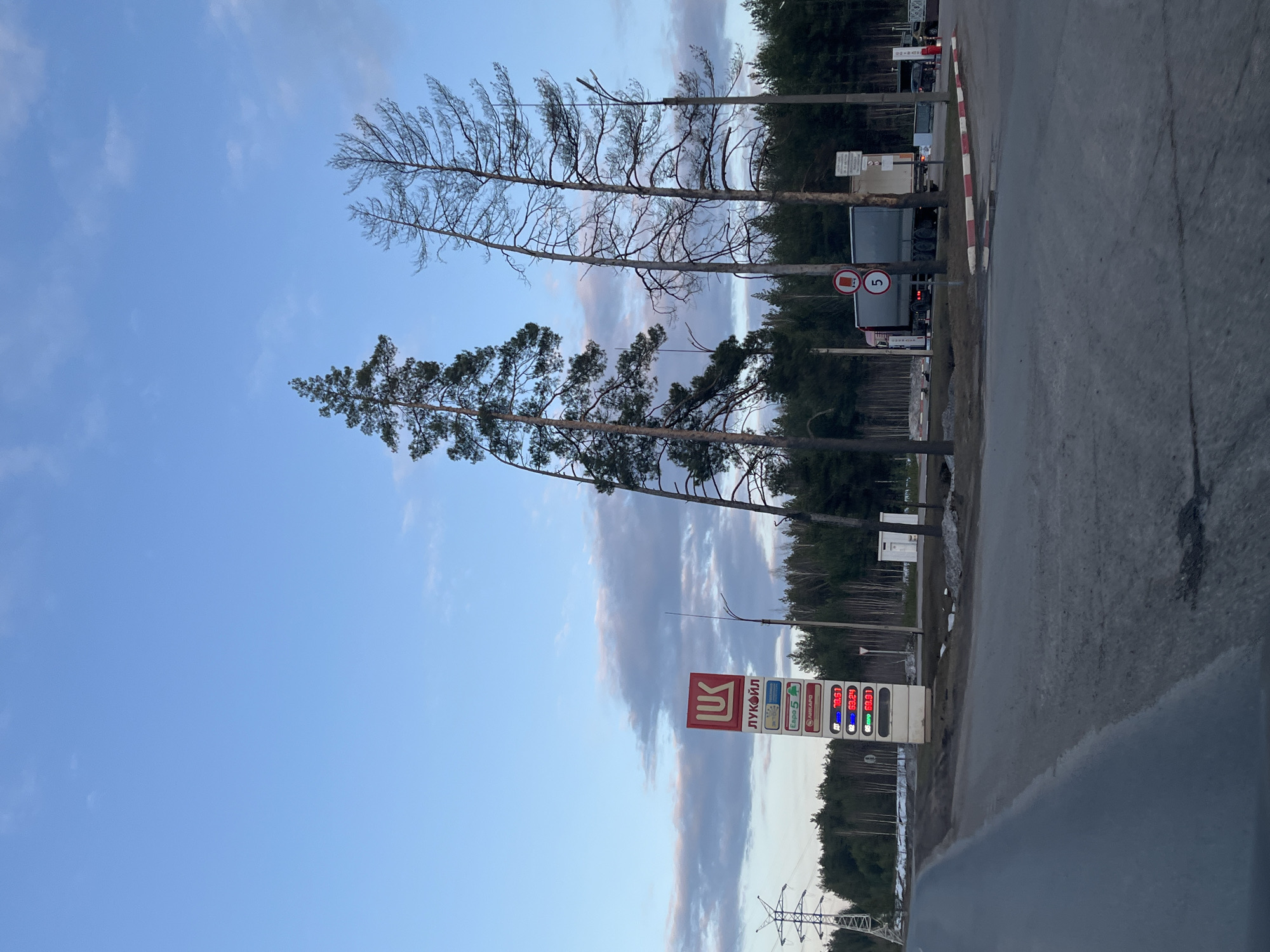The image shows a Lukoil gas station in Noviy, Russia. The gas prices are displayed on a sign, and there are trees and a cloudy sky in the background. A truck is parked nearby, and a speed limit sign is visible.