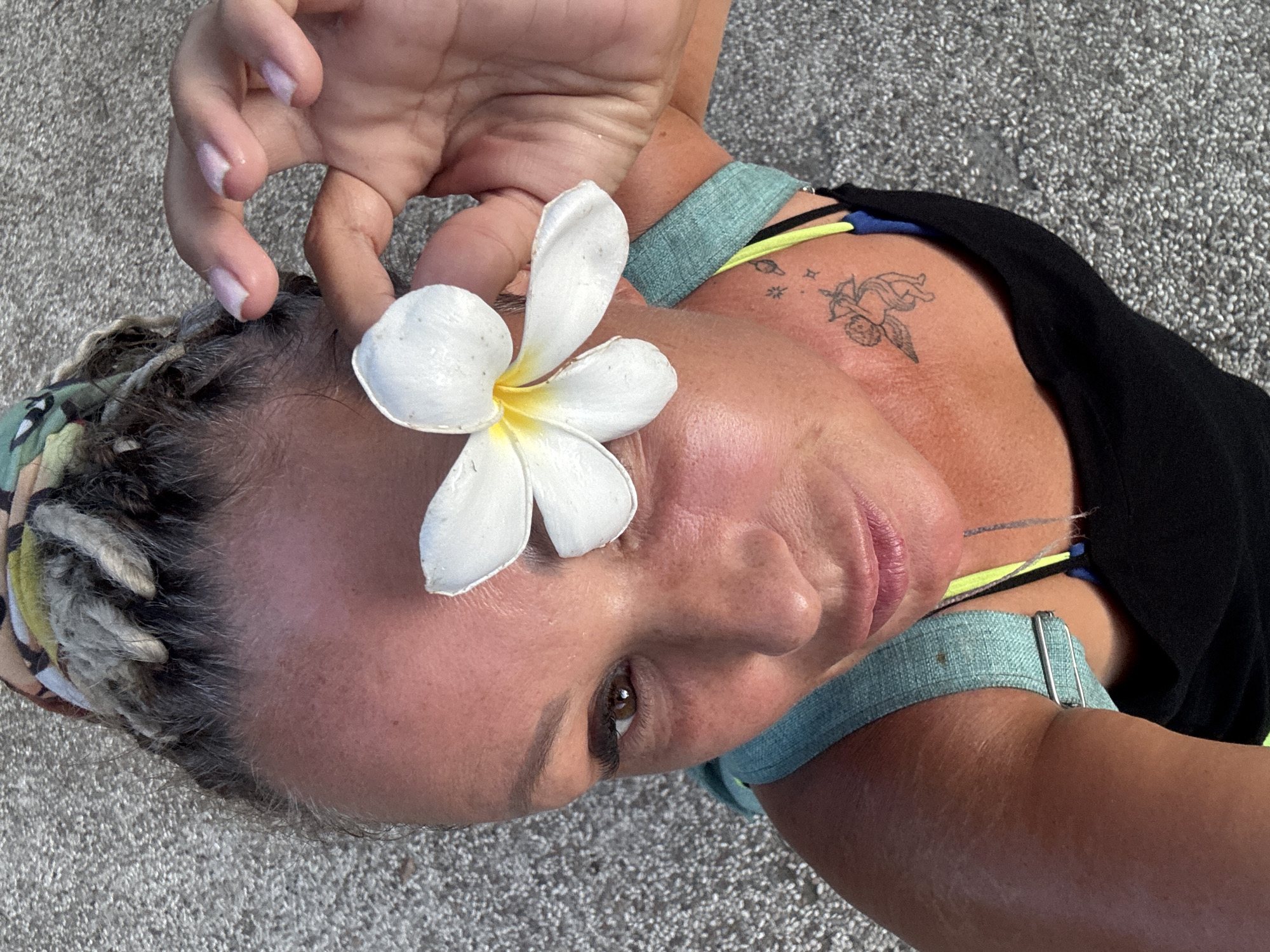 A woman with braided hair and a flower on her forehead is laying on a grey granular surface. Her right hand is positioned above the flower, while her right arm is visible in the bottom right corner, secured with a teal strap. She is wearing a black top with blue and yellow straps showing. A tattoo of an angel with a bow and arrow is visible on her shoulder. The background shows a light-grey granular texture, suggestive of sand or gravel. The woman is in Kuta Selatan, Indonesia.