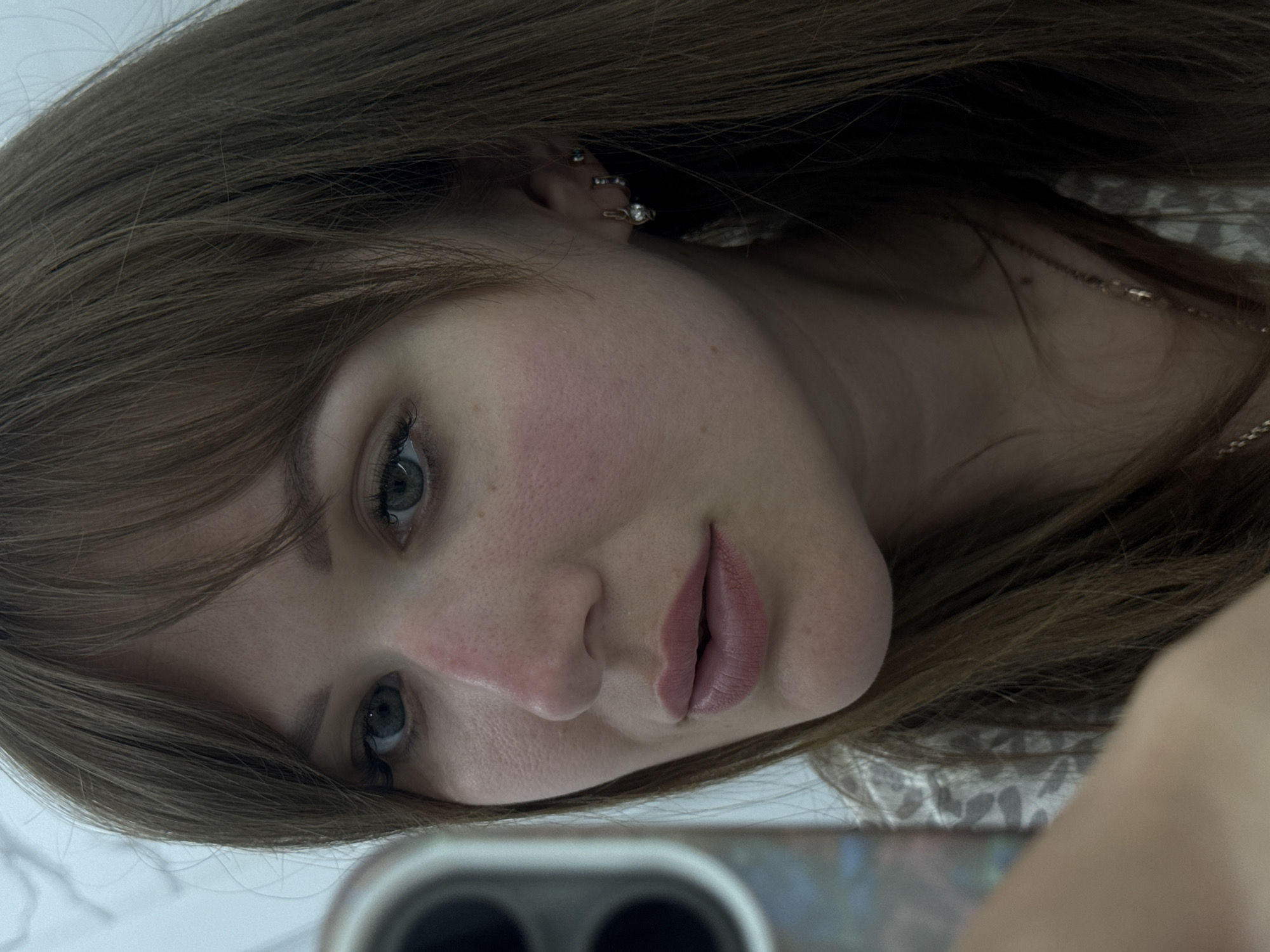 A close-up, slightly angled shot features a fair-skinned woman with brown hair partially covering her face. She has light blue eyes, prominent cheekbones with a reddish tint, and full, pale pink lips. Her hair has visible layers and a fringe partially obscuring her forehead. She has multiple ear piercings in her left ear. Part of a smartphone is visible in the lower portion of the frame, suggesting the image is a selfie. A delicate gold chain necklace is also visible. The background appears blurry and indistinct, possibly a wall or other interior surface. There is no discernible information to suggest a location in the city of Stavropol, Russia.
