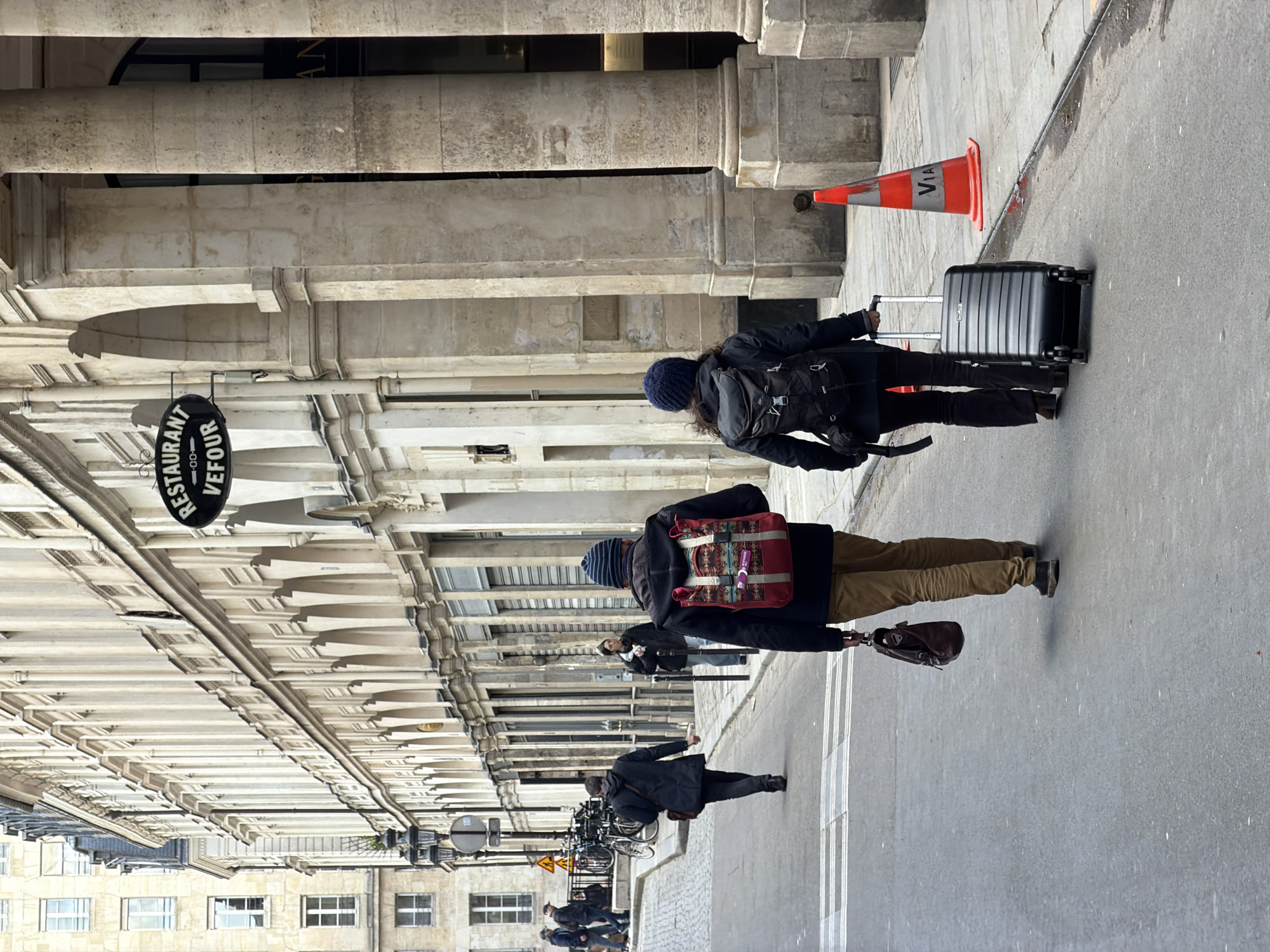 The image depicts a street scene on a cloudy day in Paris, France, specifically Rue de Beaujolais near Le Grand Véfour restaurant. In the immediate foreground, two pedestrians are observed from behind, walking away from the camera on a gray asphalt street.
The individual on the left, a male, wears a dark knit beanie, a dark jacket, tan trousers, and dark shoes. He carries a distinctive red backpack with a prominent geometric pattern and a brown leather briefcase in his right hand. The individual on the right, a female, wears a dark knit beanie, a dark jacket, dark trousers, and dark shoes, pulling a black hard-shell rolling suitcase with a silver handle. To their right, an orange traffic cone bearing the white vertical text "VIA" stands on the stone sidewalk.
Further down the street on the left, a man in a dark coat walks ahead, and a woman in a dark jacket stands on the sidewalk. A line of parked bicycles is visible near a streetlamp. The left side of the street features multiple stories of classic Parisian Haussmannian-style buildings with light-colored stone facades and numerous windows. On the right, the grand building of Le Grand Véfour restaurant is prominent, identifiable by its black oval sign with "RESTAURANT VEFOUR" in white lettering. This building showcases large fluted stone columns and ground-level arches. The street surface on the left transitions from asphalt to cobblestone, leading uphill into the distance, where additional light-colored buildings are visible under an overcast sky.