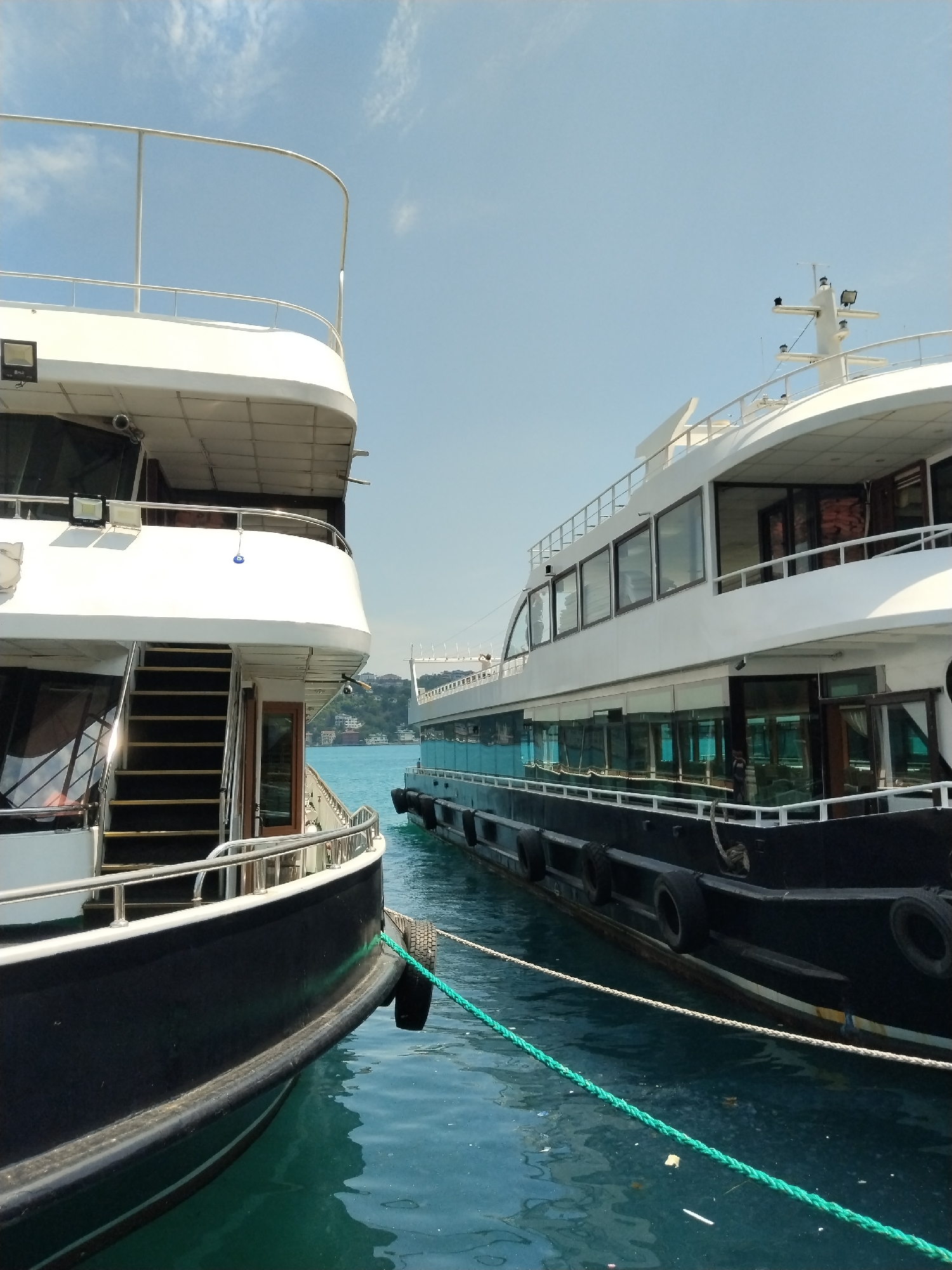 The image presents a daytime scene in Beşiktaş, Turkey, featuring two large, multi-deck boats moored side-by-side on calm, blue-green water. The sky above is a clear, bright blue with scattered wisps of white clouds, indicating sunny weather.
The boat on the left, partially in the foreground, has a sleek design with a white superstructure and a dark hull. A prominent set of stairs with dark treads and silver railings leads to an upper deck. Large windows reflect the environment, and a mounted spotlight is visible on its side. A small, blue "evil eye" charm hangs from a railing, a common cultural adornment. A thick, bright green rope secures this boat to a mooring point.
The boat on the right, further into the frame, is also a substantial vessel, likely a passenger boat or yacht. It features a white upper structure with numerous large windows extending along its length, suggesting interior viewing areas. Its dark hull is protected by several black circular fenders, which appear to be repurposed tires. A mast with various antennae or radar equipment extends from its top deck. This boat is secured by both a green and a white rope.
The water exhibits gentle ripples and reflects the clear sky and parts of the boats. In the background, beyond the boats, distant green hills dotted with residential buildings rise under the bright daylight, characteristic of a coastal city. No people are visible on or around the boats, and there is no discernible text visible. The scene suggests a moment of rest or preparation for activity at a dock or pier.