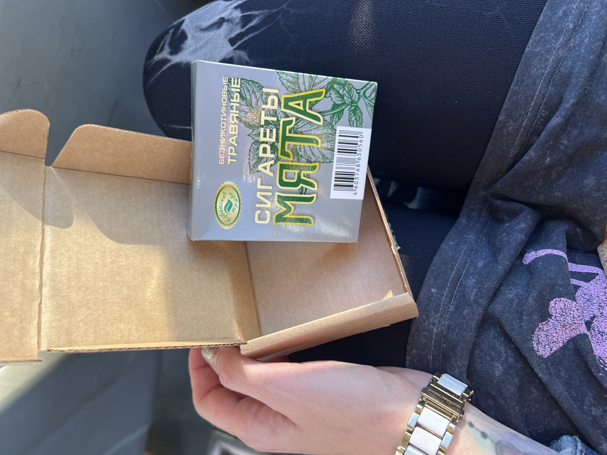 A person's hands are visible in the foreground, holding open a small, light brown cardboard box. Inside the box, a pack of cigarettes with a mint design is placed upright. The pack is silver with green text and an illustration of mint leaves. It appears to be a sunny day, as indicated by the shadows and the bright lighting. The person is wearing a watch with a white and gold band on their left wrist. In the background, dark blue fabric with a mottled texture, possibly clothing, is visible. The scene seems to be outdoors or in a well-lit indoor space, given the strong sunlight. The text on the cigarette pack reads "СИГАРЕТЫ МЯТА" (Cigarettes Mint) and "БЕЗНИКОТИНОВЫЕ ТРАВЯНЫЕ" (Nicotine-Free Herbal). There is also a circular logo with green text and an image of leaves, and a barcode with the numbers "4 603768 650560". The overall impression is casual, with the person examining or presenting the contents of the box.