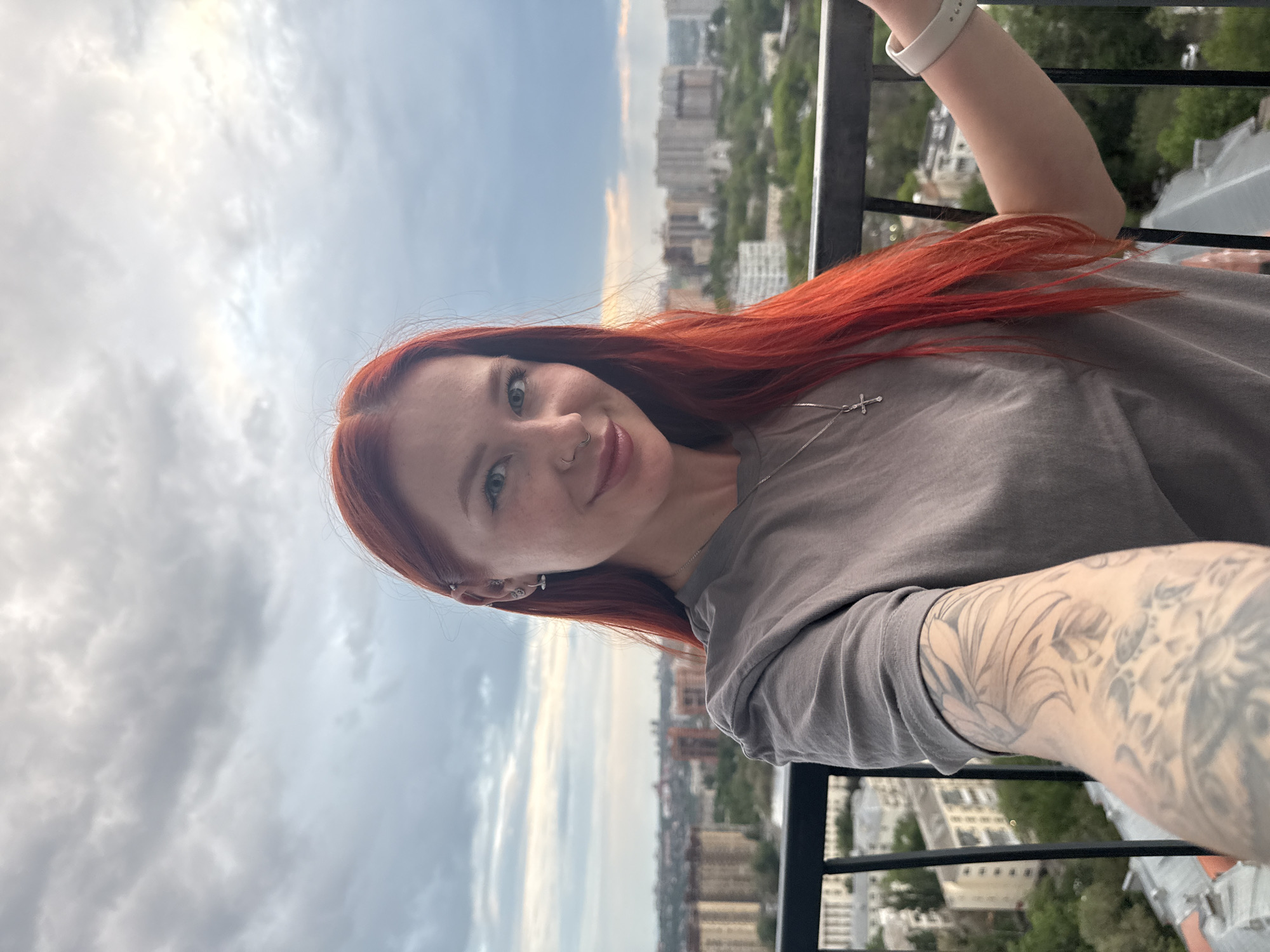A woman with red hair is standing on a balcony overlooking the city of Rostov-on-Don, Russia. She is wearing a gray t-shirt and a silver necklace with a cross pendant. She has a nose ring and multiple earrings on her left ear, and a tattoo on her left arm. She is holding a white watch on her left wrist. The sky is cloudy, and the cityscape is visible in the background. The balcony has a black railing.