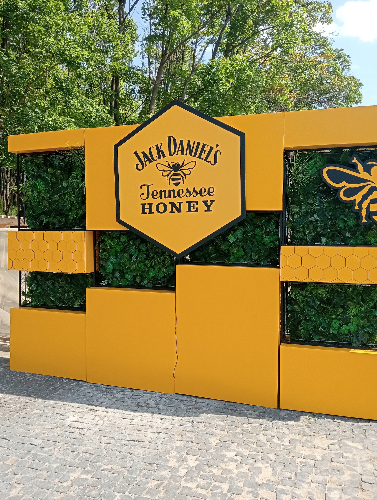 The image displays a bright yellow, modern outdoor structure designed to resemble a honeycomb. The structure features geometric shapes and incorporates sections filled with lush green artificial foliage. Dominating the center of the structure is a large hexagonal sign with the "Jack Daniel's Tennessee Honey" logo, complete with a bee graphic. To the right of the logo, another large bee graphic is visible, also in black and yellow. The structure is set against a backdrop of mature green trees and a clear blue sky. In the foreground, a cobblestone pathway is visible, casting a shadow from the structure. The overall scene appears to be an outdoor promotional display or event setup, possibly in a park or public area, given the natural surroundings. The location is identified as Obariv, Ukraine. There are no people visible in the image.